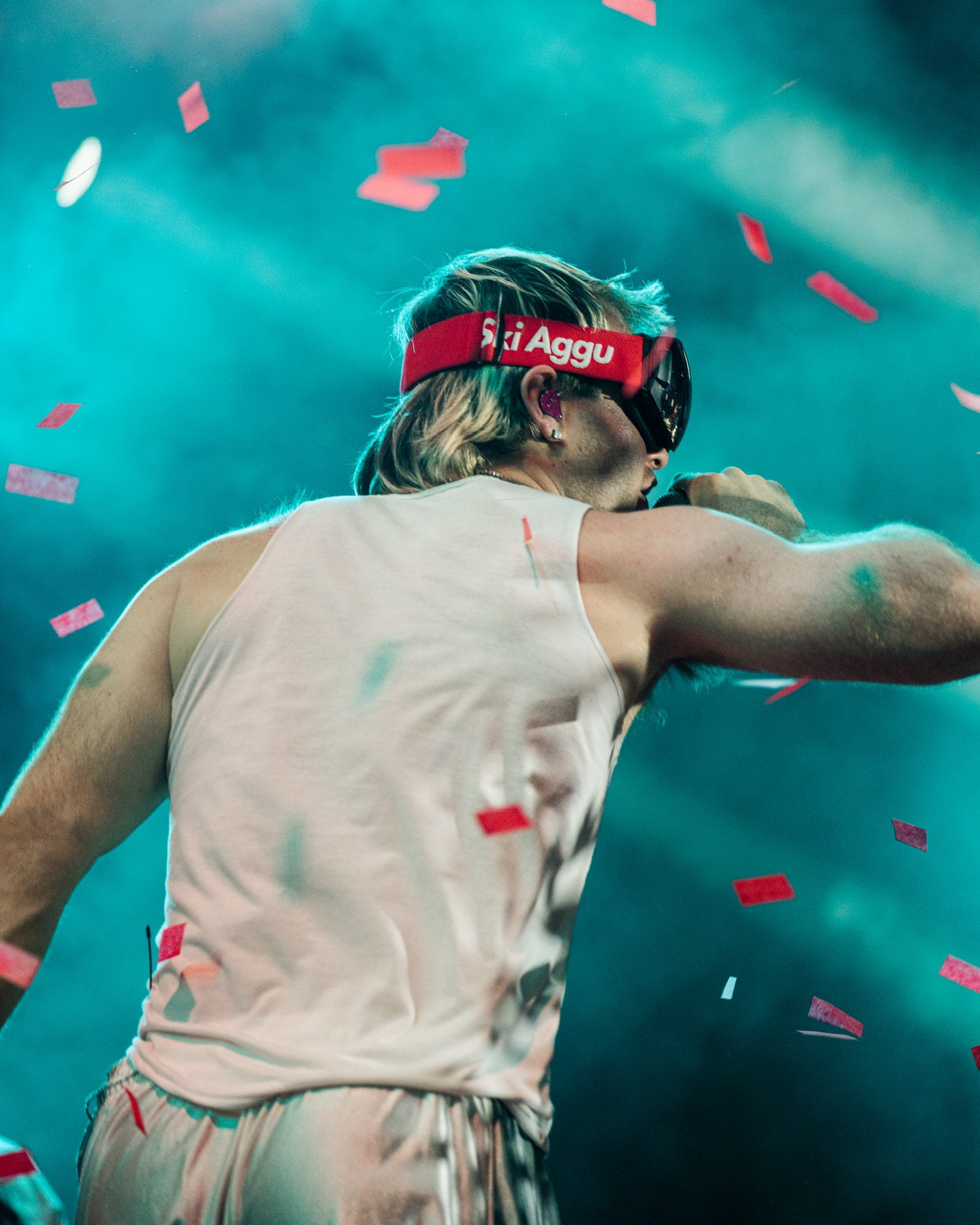 Ein männlicher Musiker während eines Konzerts, trägt eine Virtual-Reality-Brille mit der Aufschrift 'SkI Aggu' und eine weiße Tanktop, während rote Konfetti im Hintergrund fällt.