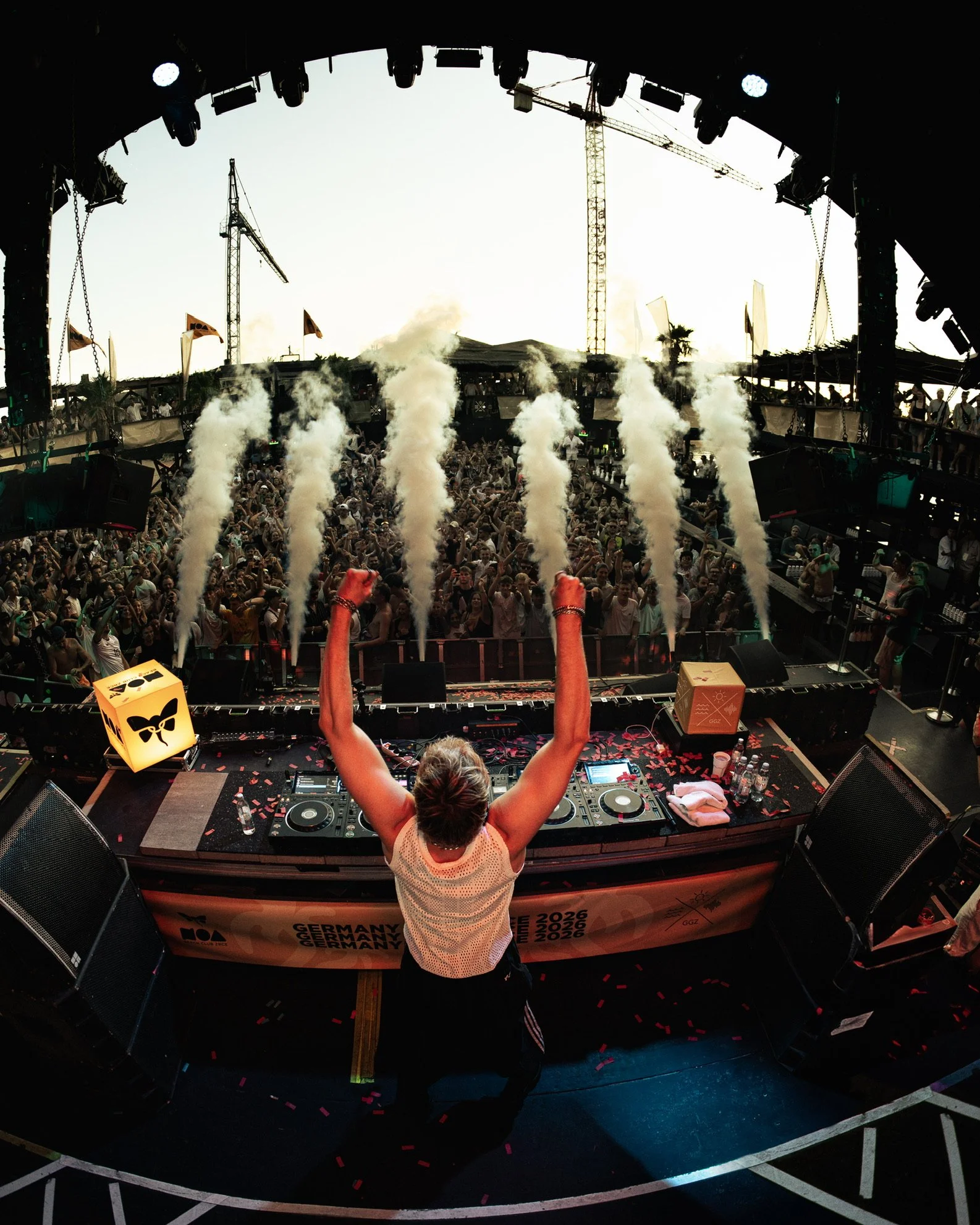 DJ beim Auflegen auf einer Bühne vor großer Menschenmenge während eines Festivals, mit Rauch- und Lichteffekten im Hintergrund.