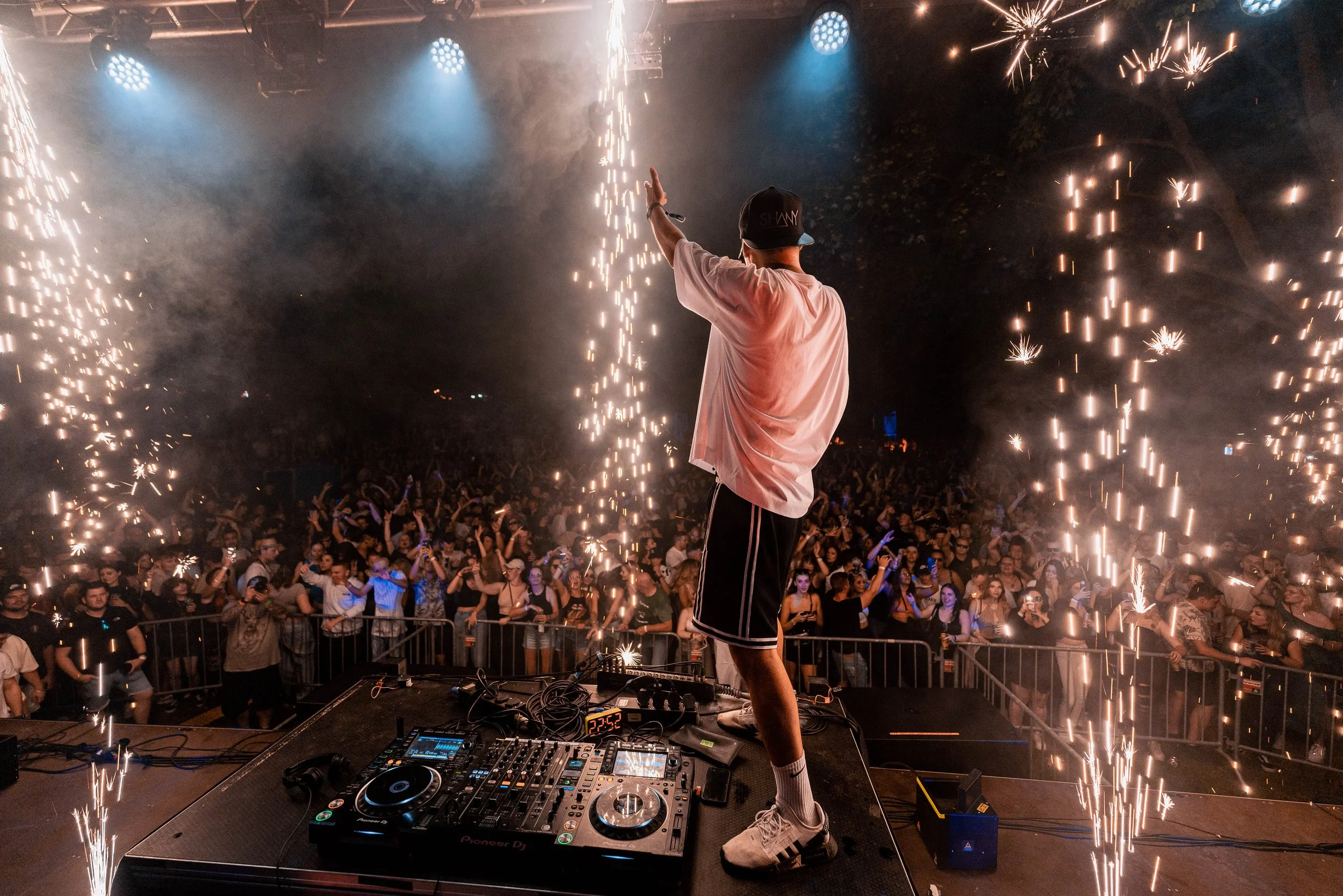 Ein DJ auf einer Bühne während eines Live-Auftritts mit Feuerwerks- und Funkeneffekten, vor einer großen Menschenmenge bei Nacht.