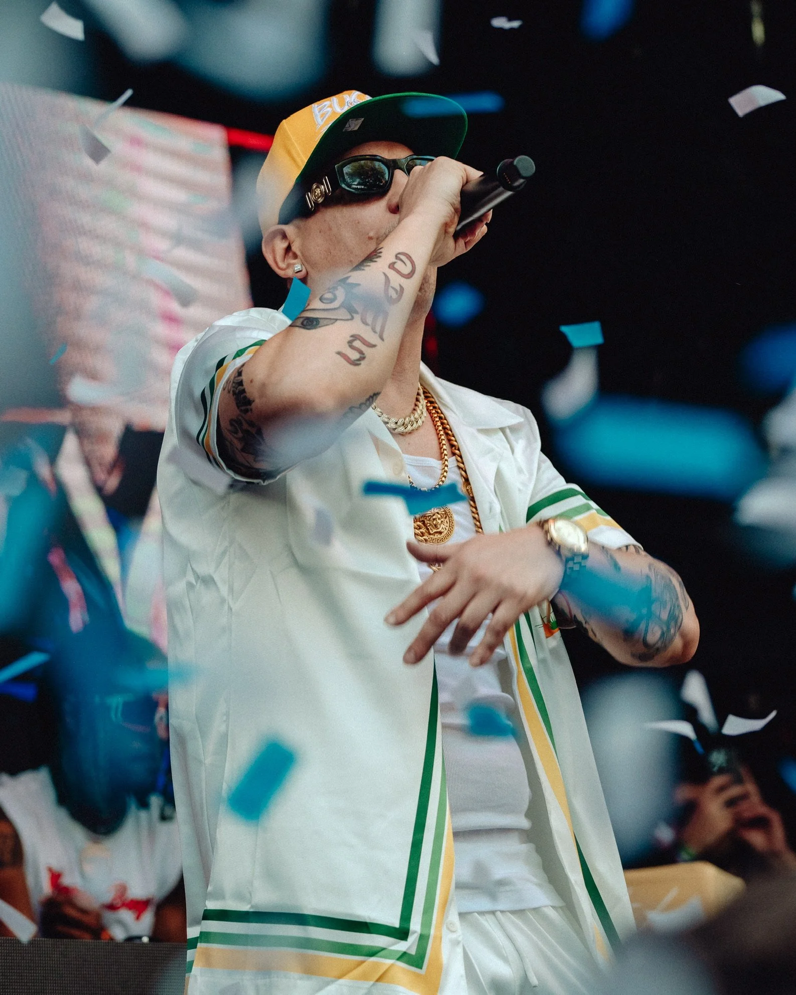 Ein Mann mit Tattoos, Sonnenbrille und Basecap, der auf einer Bühne singt und greifbare Konfettiteilchen fallen lässt.