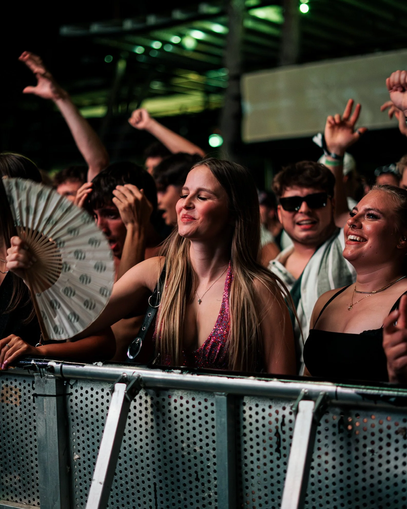 Junge Frauen und Männer feiern auf einem Konzert oder Festival, tanzen, lachen und genießen die Atmosphäre, mit Metallabsperrung im Vordergrund.