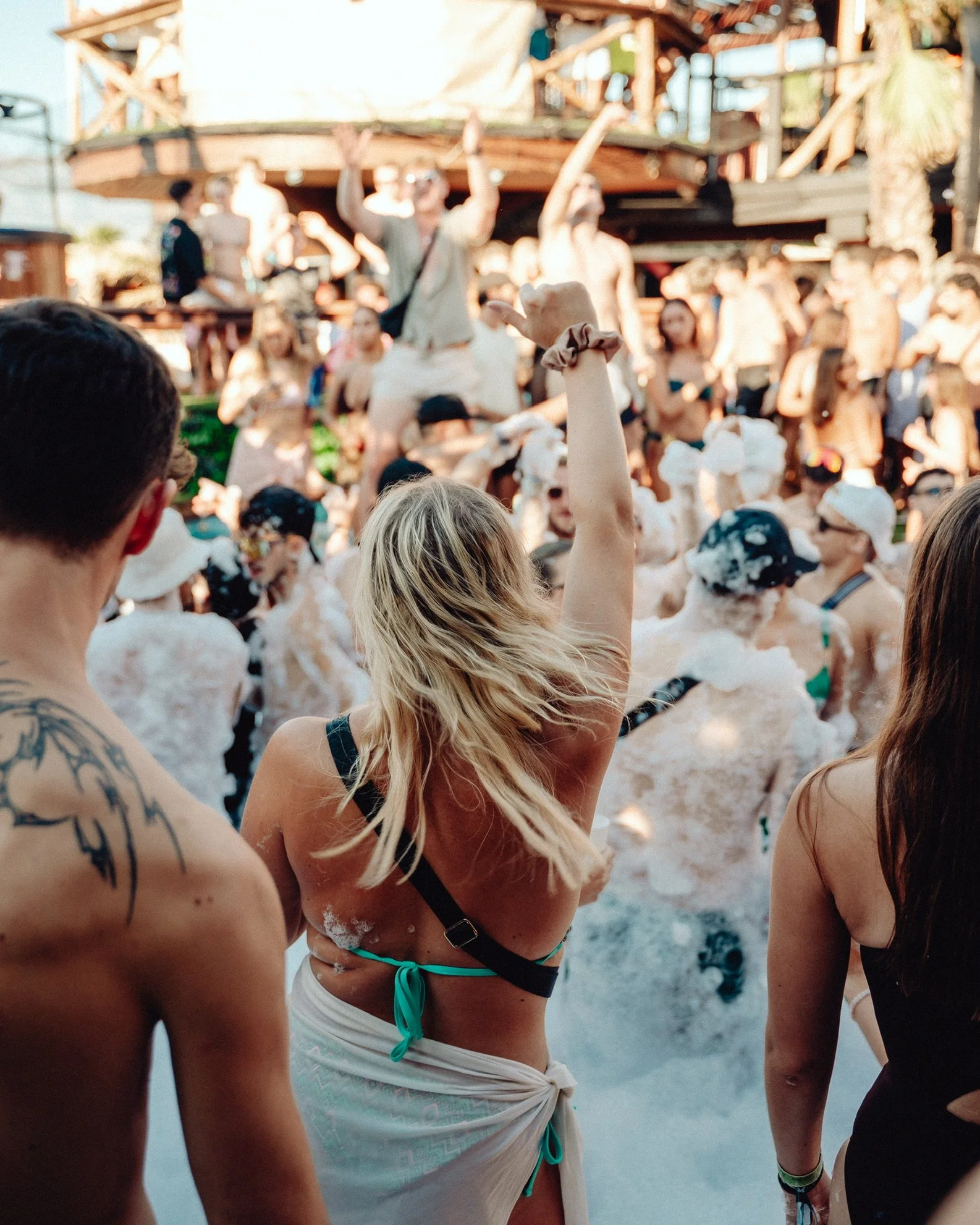 Menschen feiern auf einer Party im Wasser mit Seifenblasen, im Hintergrund eine Holzterrasse und weitere Partygänger.