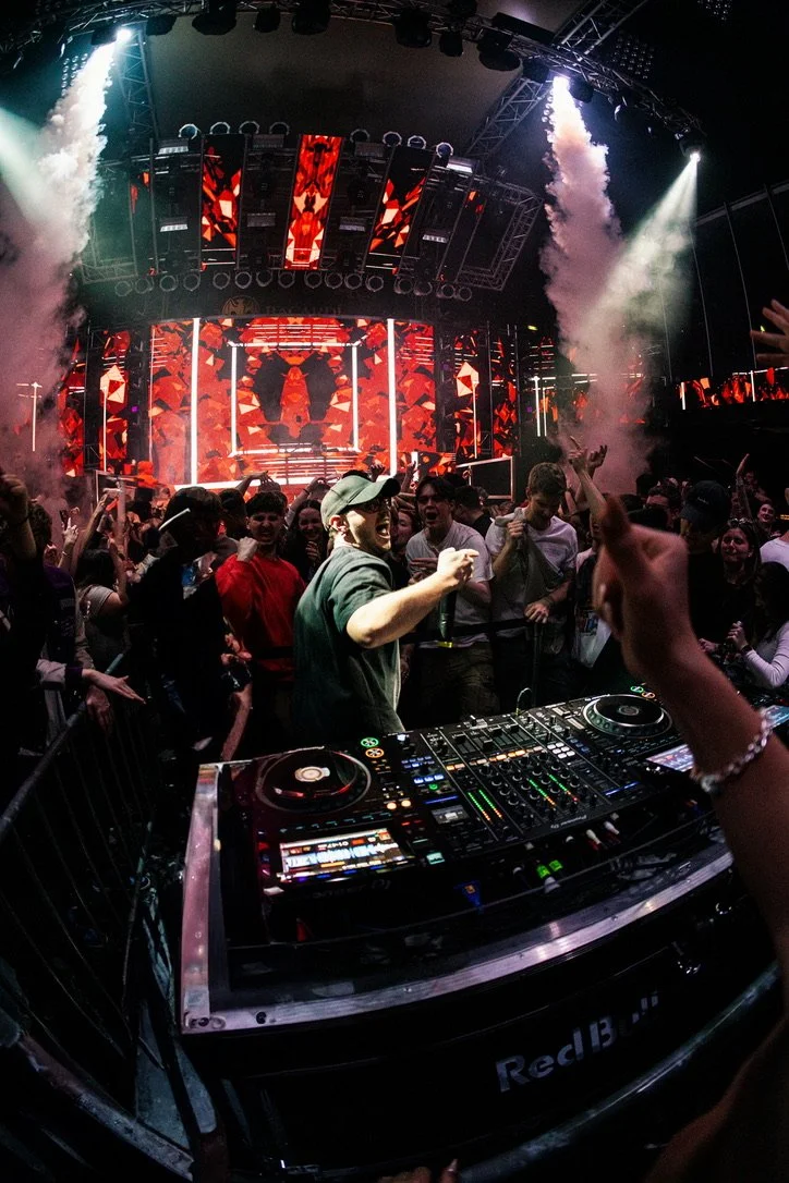Menschen feiern in einer Diskothek mit einer großen, hell erleuchteten Bühne und einem DJ-Setup.