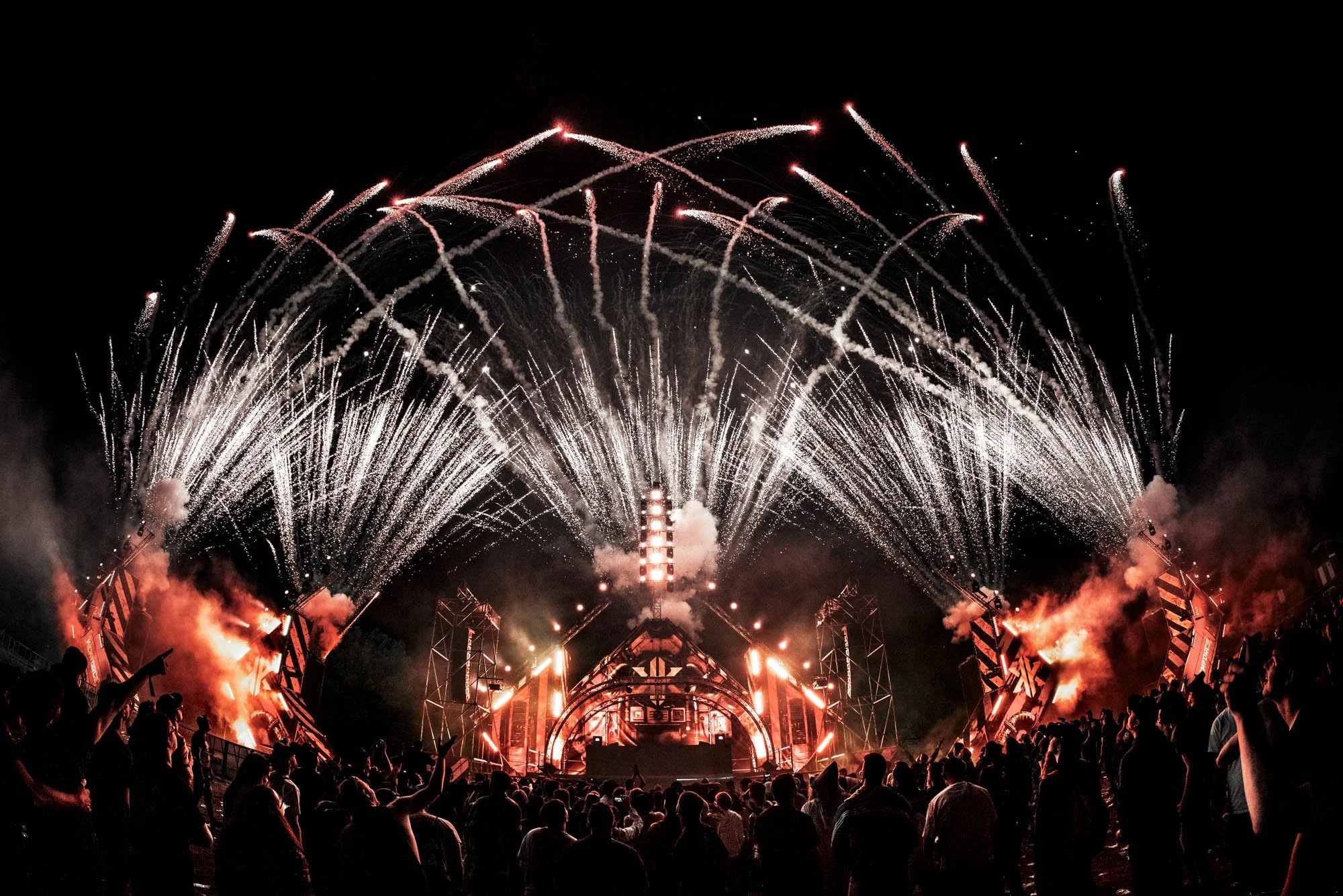 Menschen feiern auf einem Konzert im Freien bei Nacht, während Feuerwerk am Himmel explodiert.