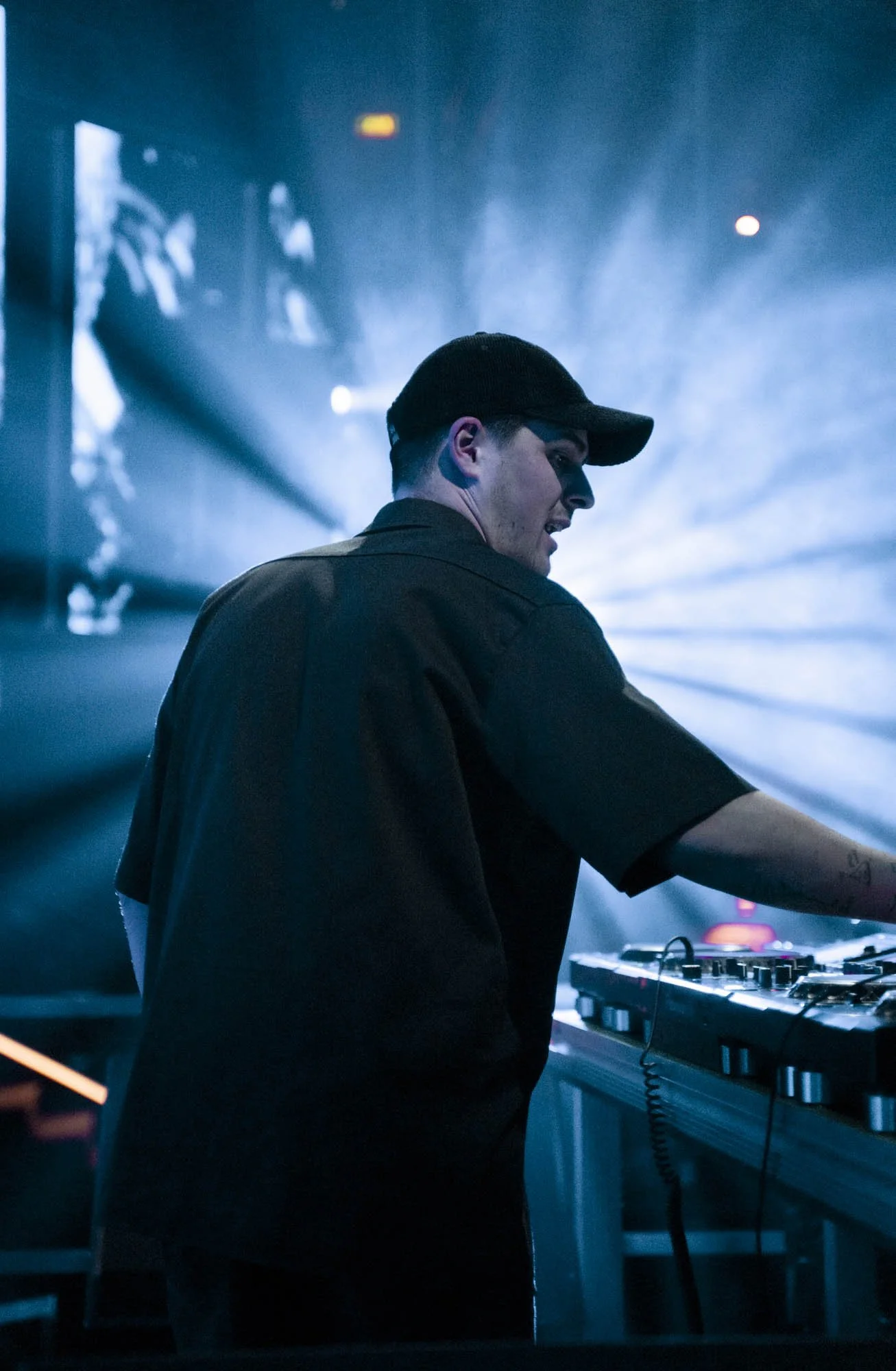 Ein junger Mann in dunkler Kleidung und Baseballkappe, der an einem DJ-Pult in einem Club oder Konzertsaal steht, umgeben von blauen Lichteffekten.