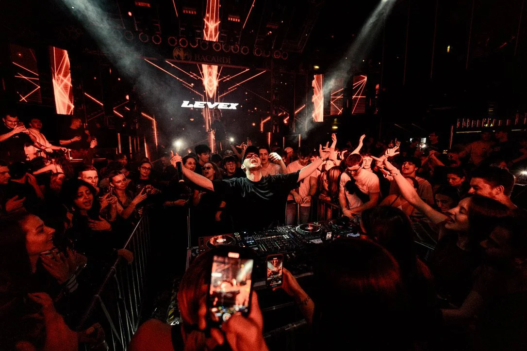 Menschen feiern bei einem DJ-Set in einem dunklen Club mit roten Licht- und LED-Displays im Hintergrund.