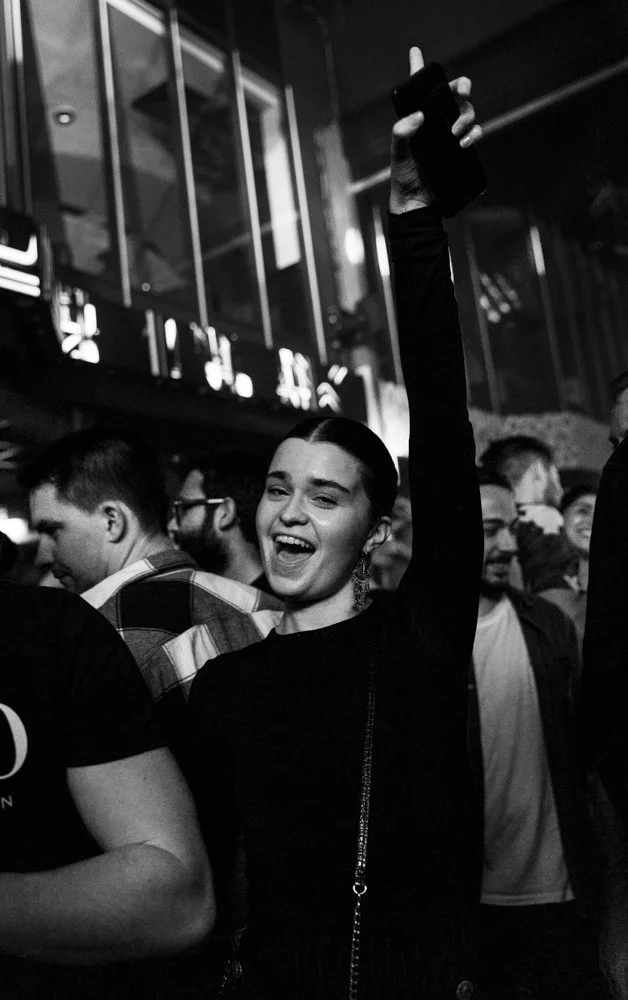 Eine lachende junge Frau in dunkler Kleidung, die auf einer Party mit vielen Menschen im Hintergrund tanzt und ein Handy in die Luft hält.