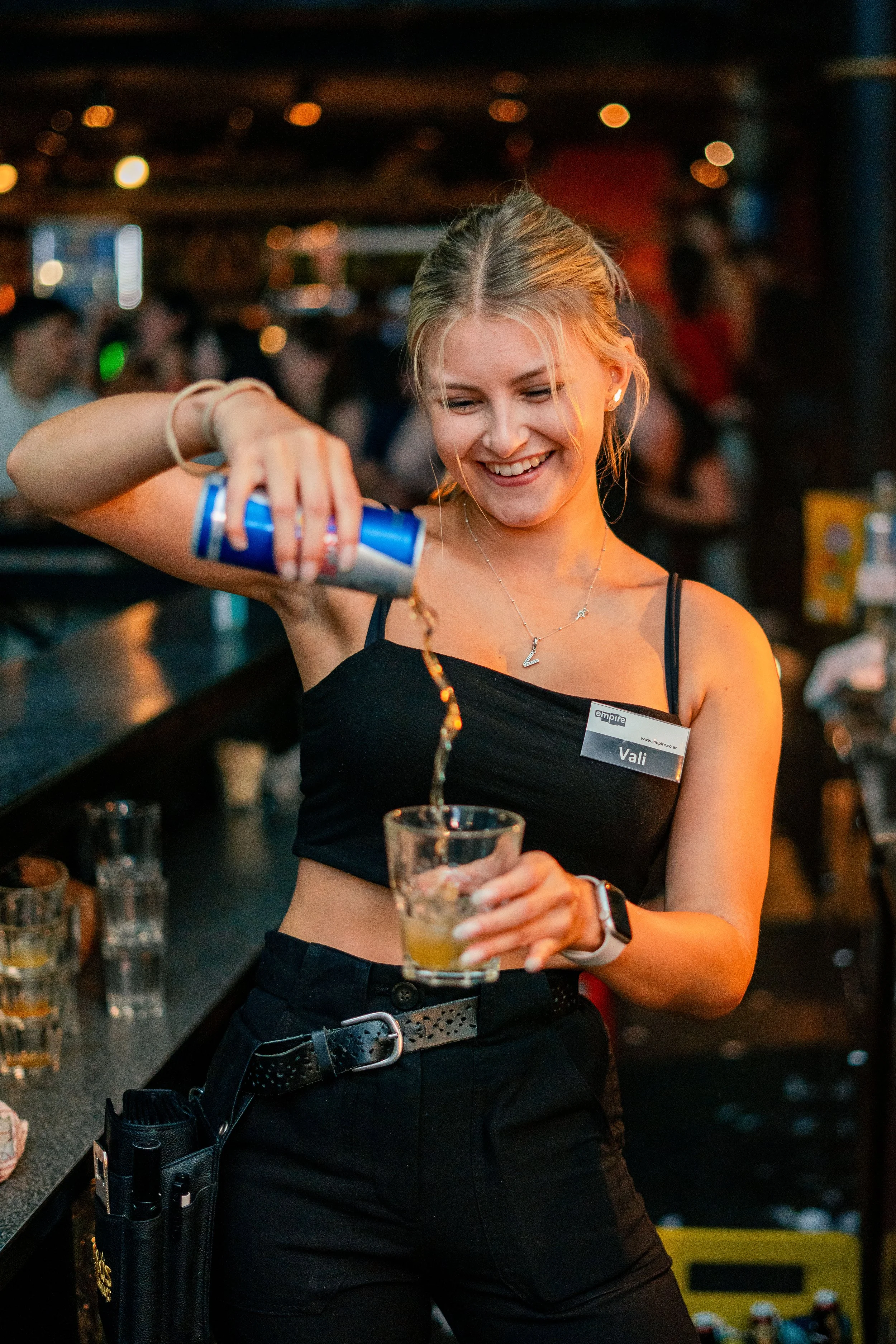 Eine junge Frau mit Namensschild beim Mixen eines Getränks in einer Bar, lachend, mit dunklem Top und dunklen Hosen.