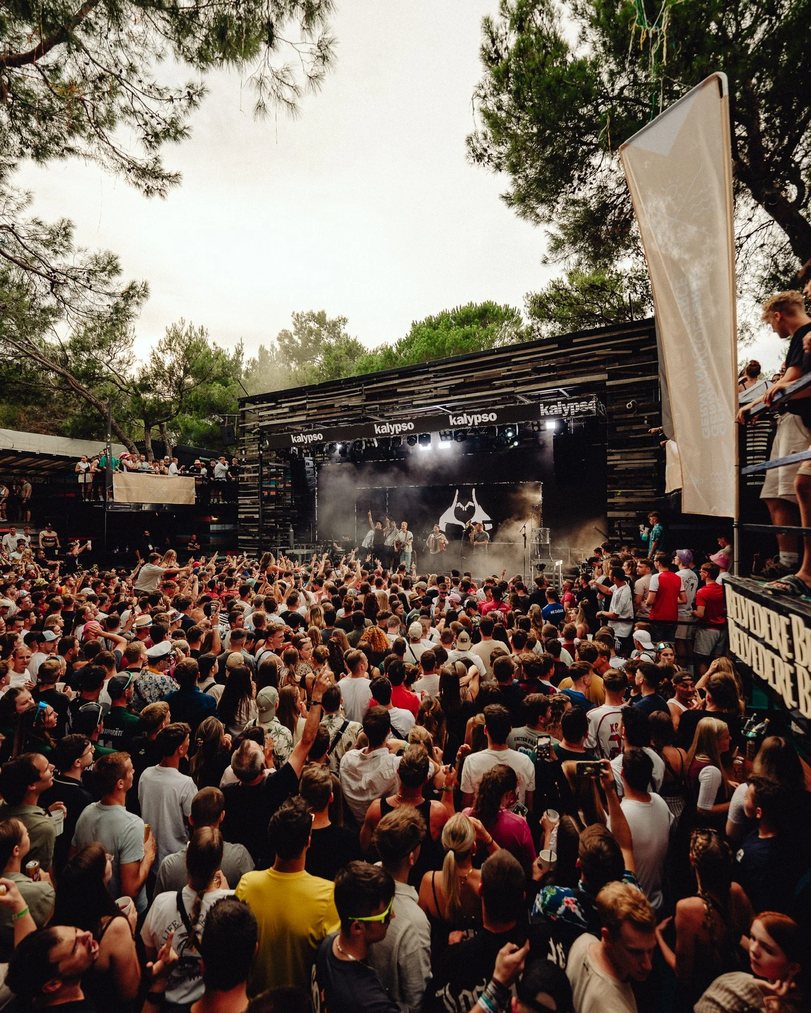 Ein Konzert im Freien mit einer großen Menschenmenge vor einer Bühne, auf der eine Band spielt, umgeben von Bäumen bei Tageslicht.