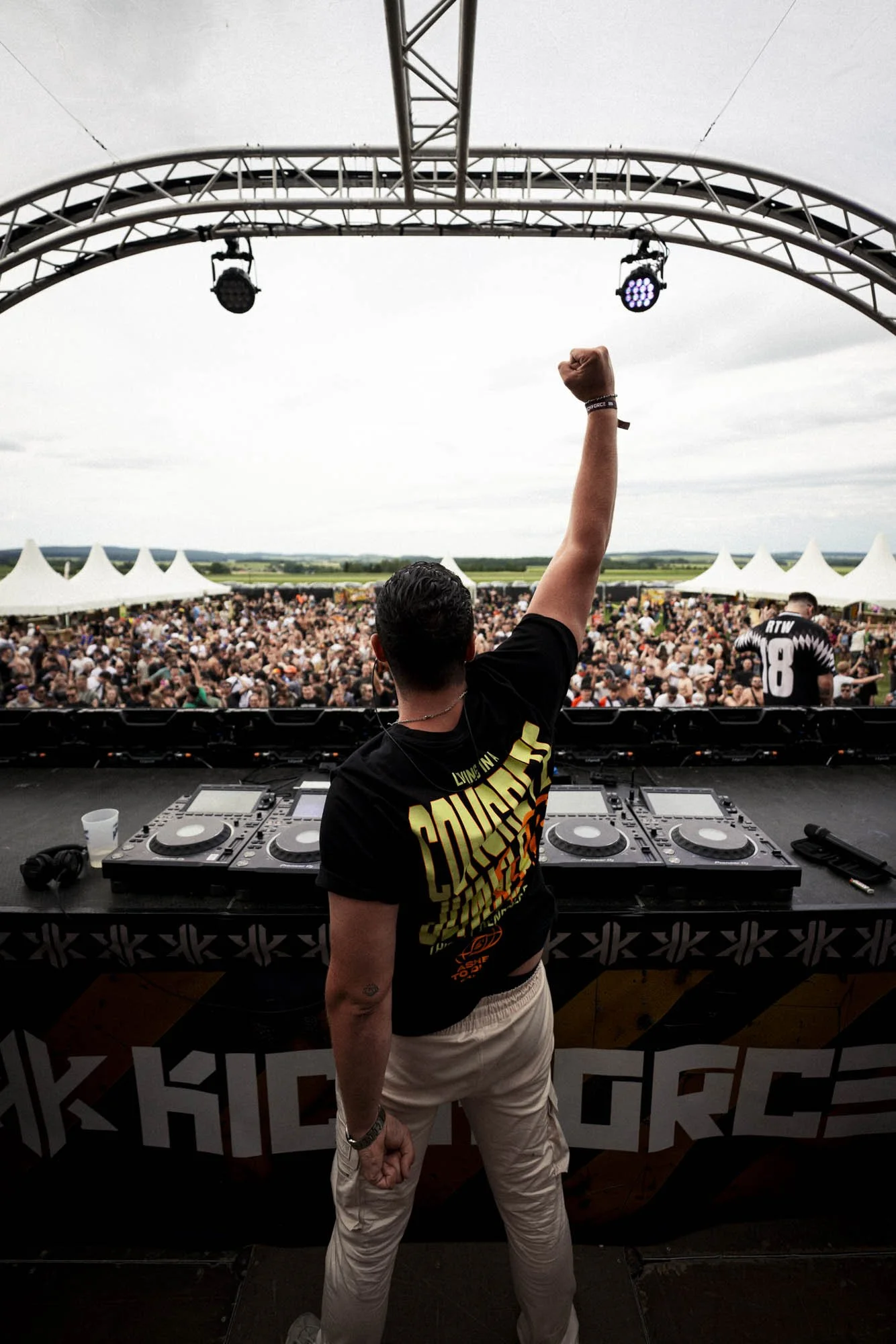 DJ auf einer Bühne hebt die Faust vor einem großen Publikum bei einem Open-Air-Konzert, mit weißen Zelten im Hintergrund und einem grauen Himmel.