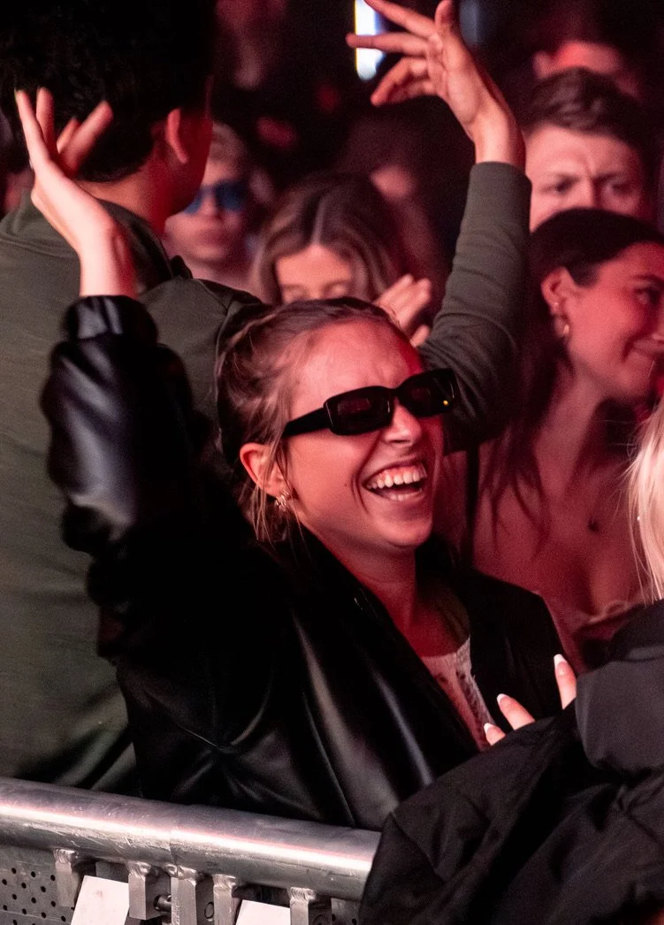 Junge Frau mit Sonnenbrille, lachend auf einem Konzert oder Festival, umgeben von anderen Menschen.