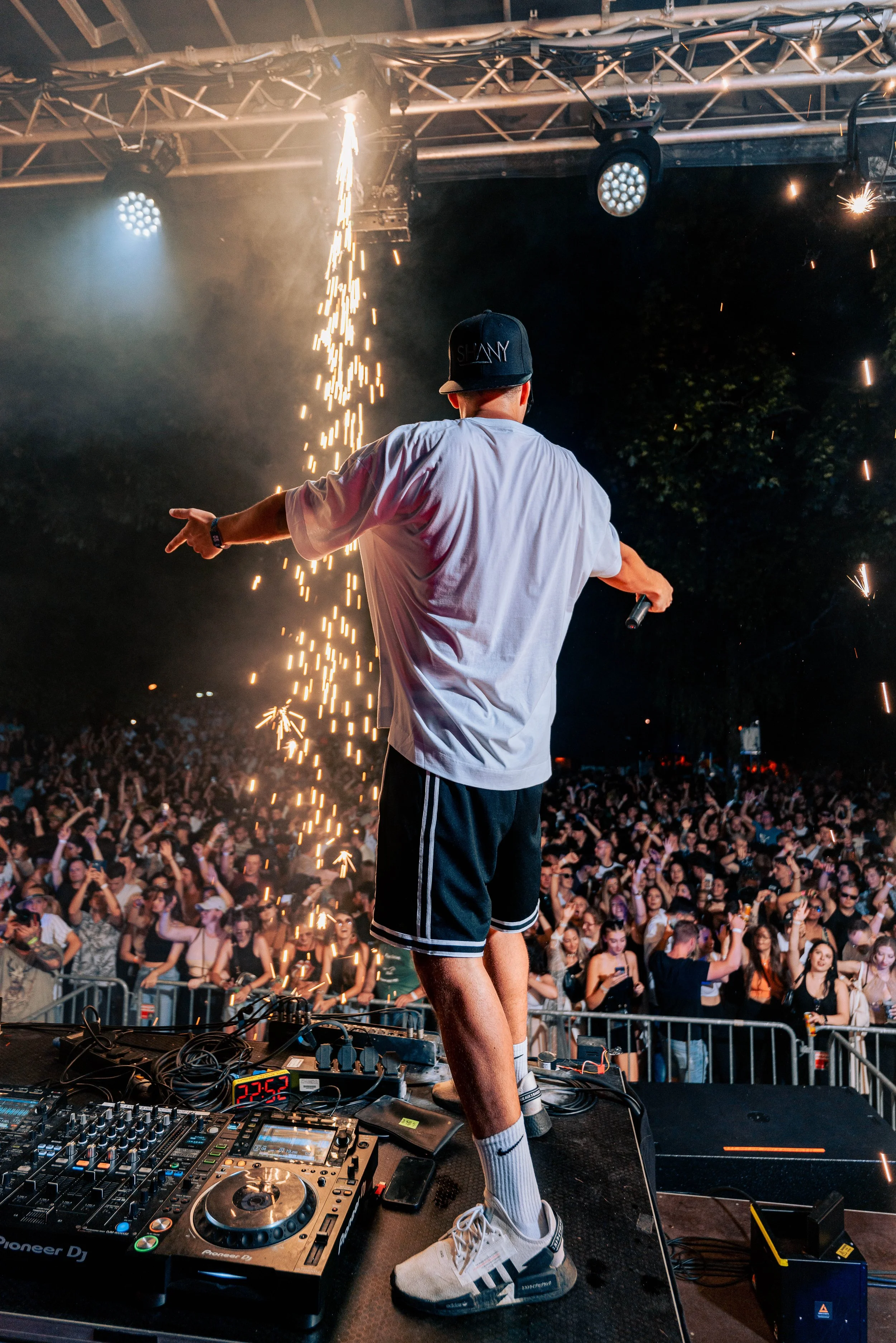 DJ auf einer Bühne bei einem Nachtfestival, hinter ihm ein großer Menschenmenge, die Spaß hat, während Feuerwerks- oder Funken-Show im Hintergrund gesprüht werden.
