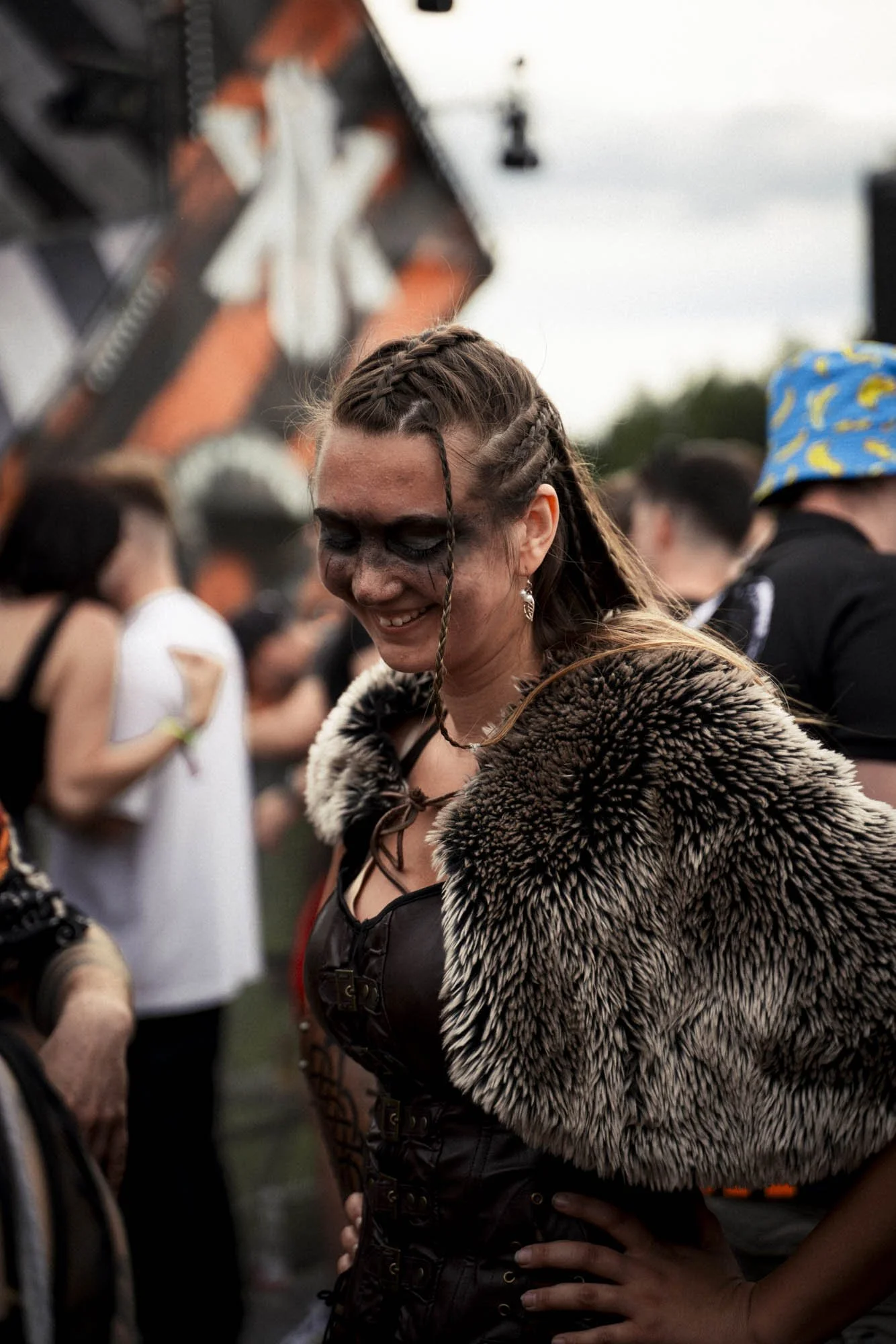 Eine lachende Frau mit gestyltem Haar und auffälligem Make-up trägt ein Fellcape auf einer Musik- oder Festivalveranstaltung, umgeben von anderen Besuchern.