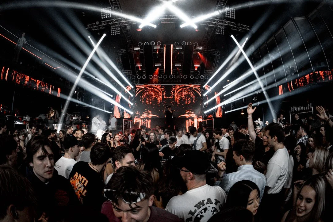 Menschen tanzen und feiern in einer Club-Umgebung mit einer Bühne, bunten Lichtern und einem DJ oder Performer im Zentrum.