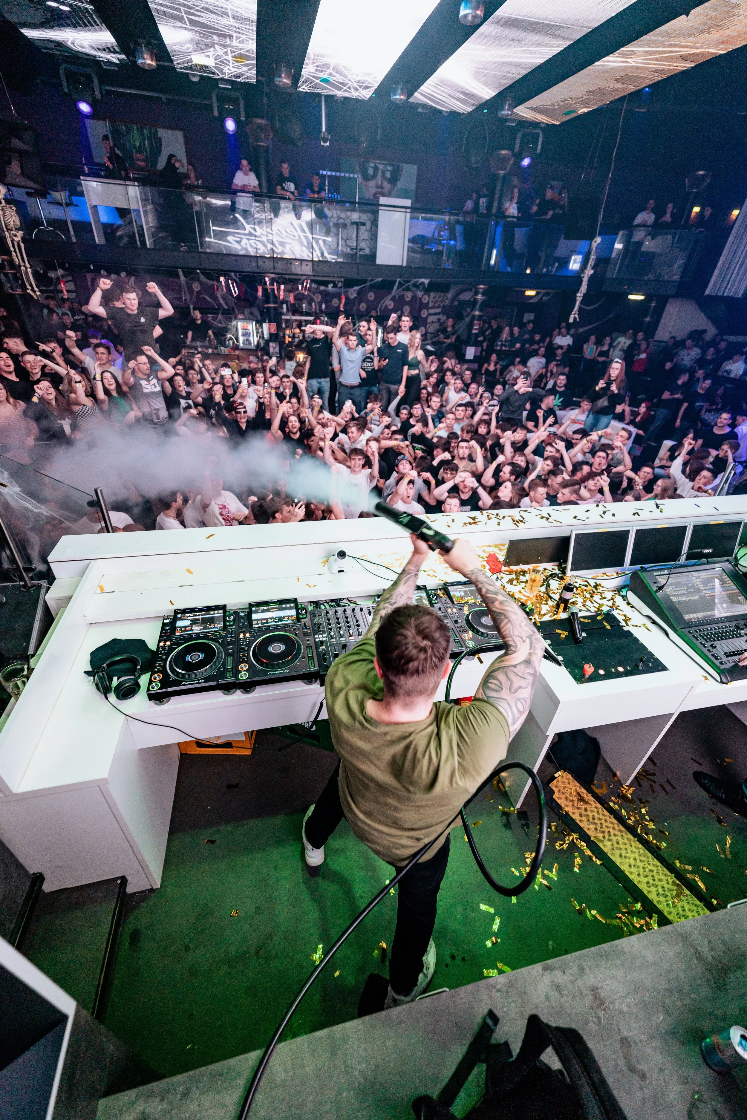 Ein DJ steht hinter einem Mischpult auf einem DJ-Pult, während vor ihm eine ausgelassene Menschenmenge in einem Club tanzt, mit Konfetti auf dem Boden und Nebel in der Luft.