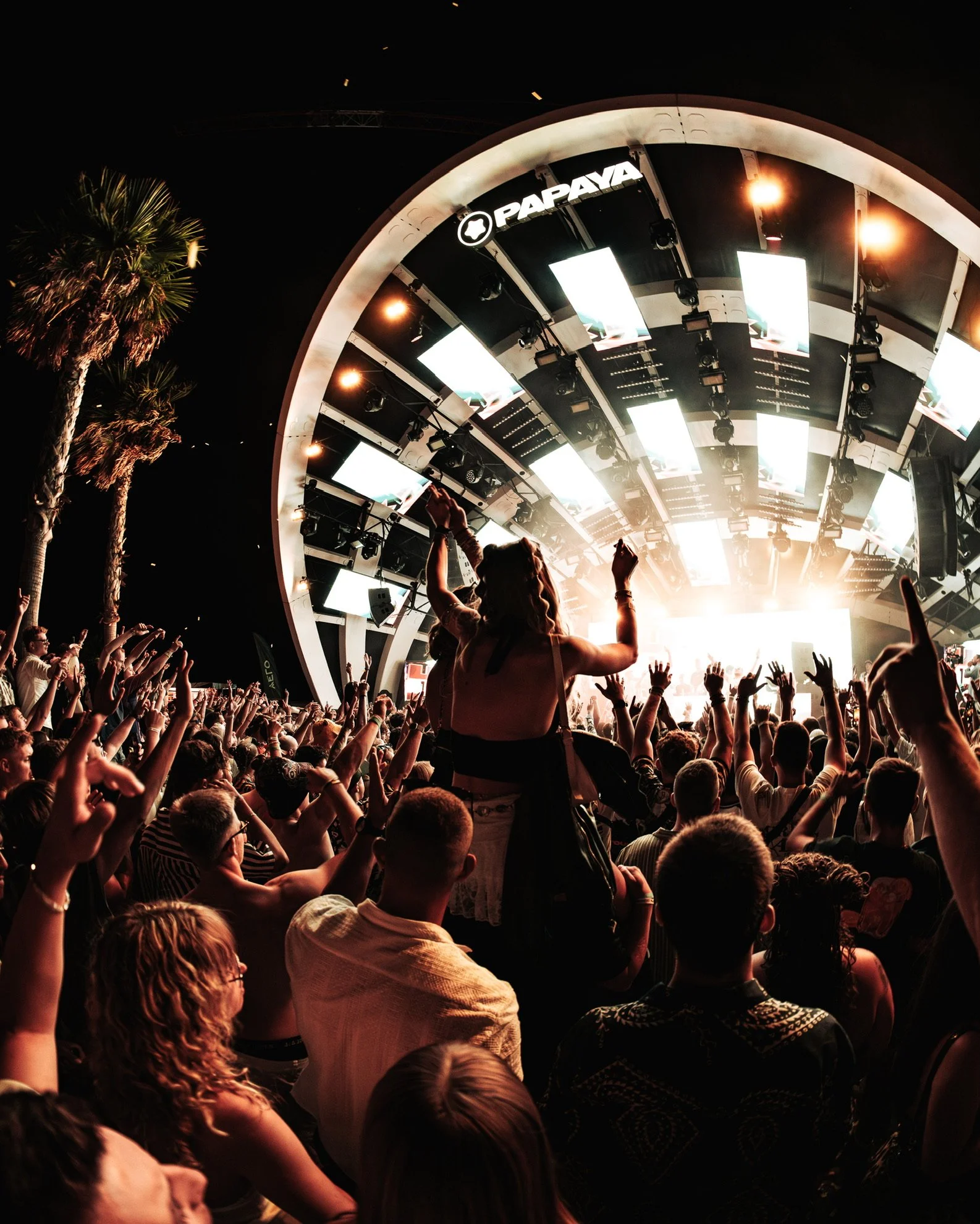 Menschen tanzen und feiern bei einem Musikfestival vor einer großen Bühne mit Scheinwerfern und Bildschirmen bei Nacht, Palmen im Hintergrund.