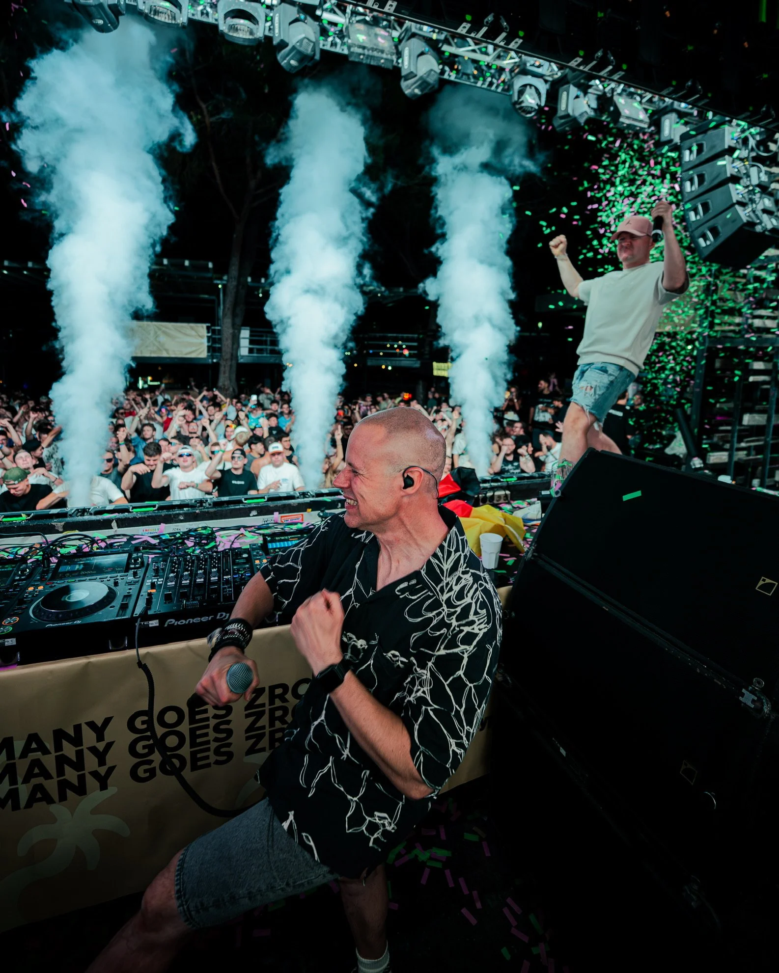 DJ und ein Tänzer feiern auf einer Party, im Hintergrund ein großes Publikum, Rauch- und Konfettispuren in der Luft.