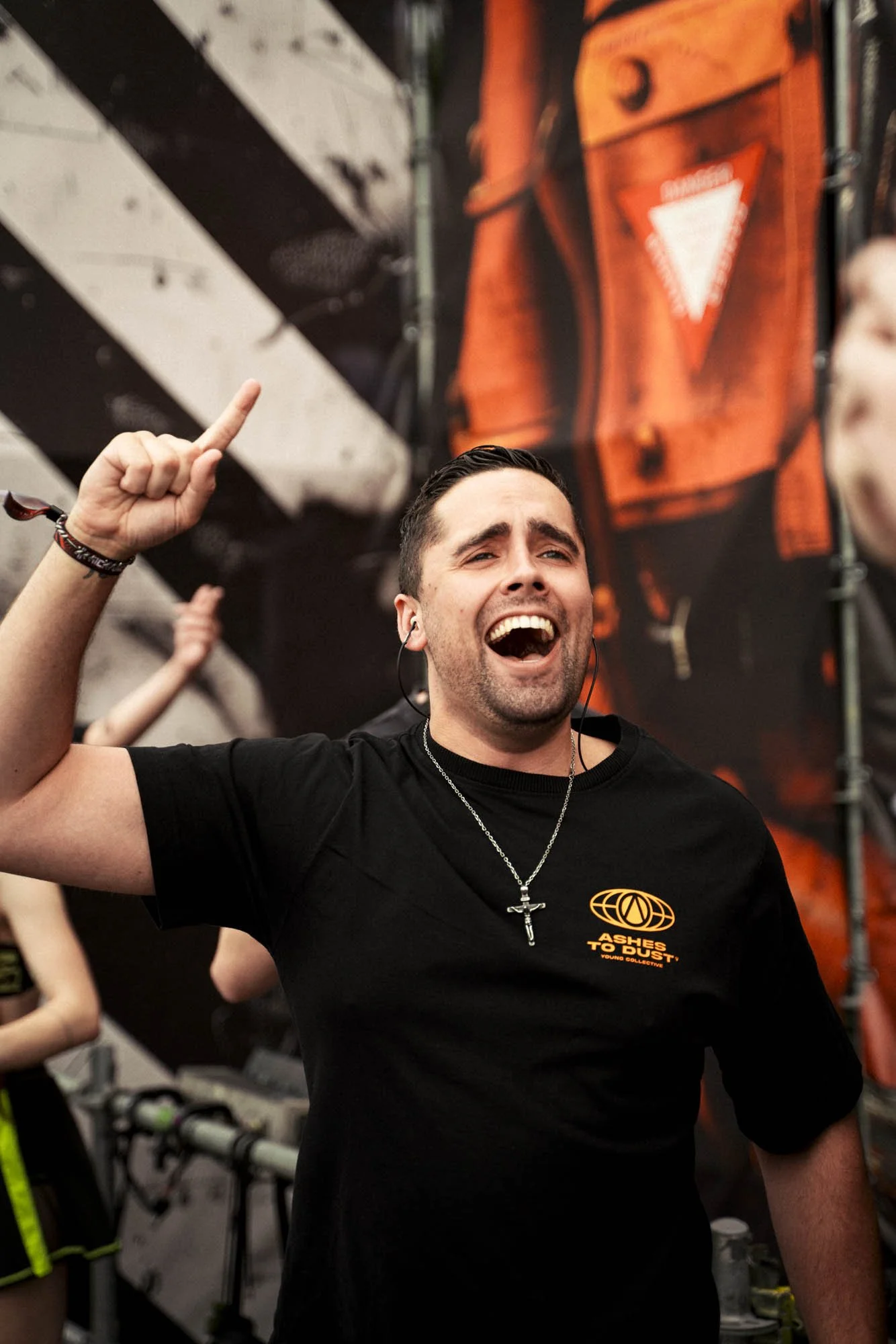 Ein lachender Mann mit schwarzen Haaren, schwarzem T-Shirt mit Gelb-Logo und Kreuz-Halskette, der bei einem Konzert oder Festival Freude zeigt.