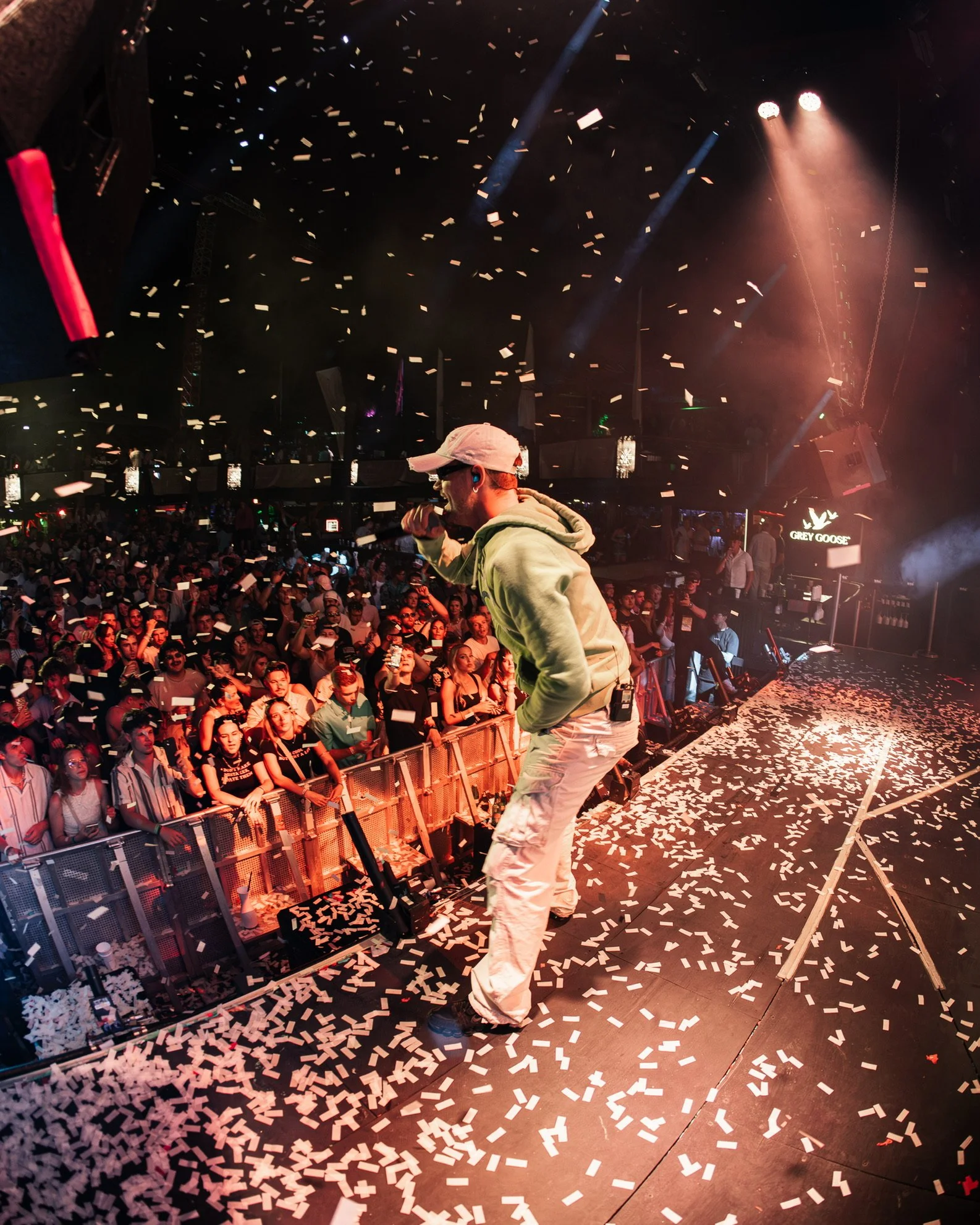Ein Performer auf der Bühne in einem Club, umgeben von einem Publikum, mit Konfetti in der Luft und auf dem Boden, während bunte Lichter den Raum beleuchten.
