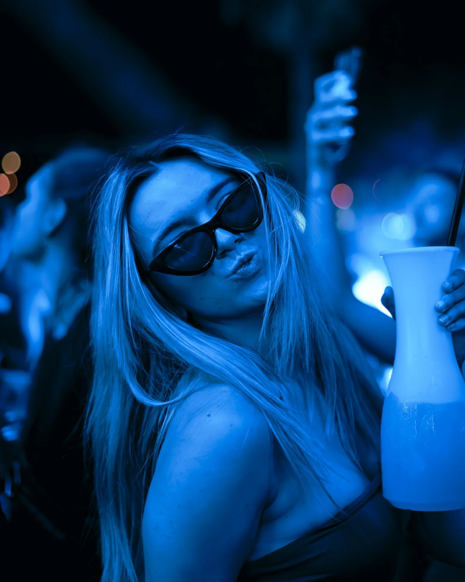 Eine junge Frau mit langen blonden Haaren trägt dunkle Sonnenbrille und macht ein Kussgesicht, während sie in einer nächtlichen Umgebung mit blauen Lichtverhältnissen eine Vase hält.