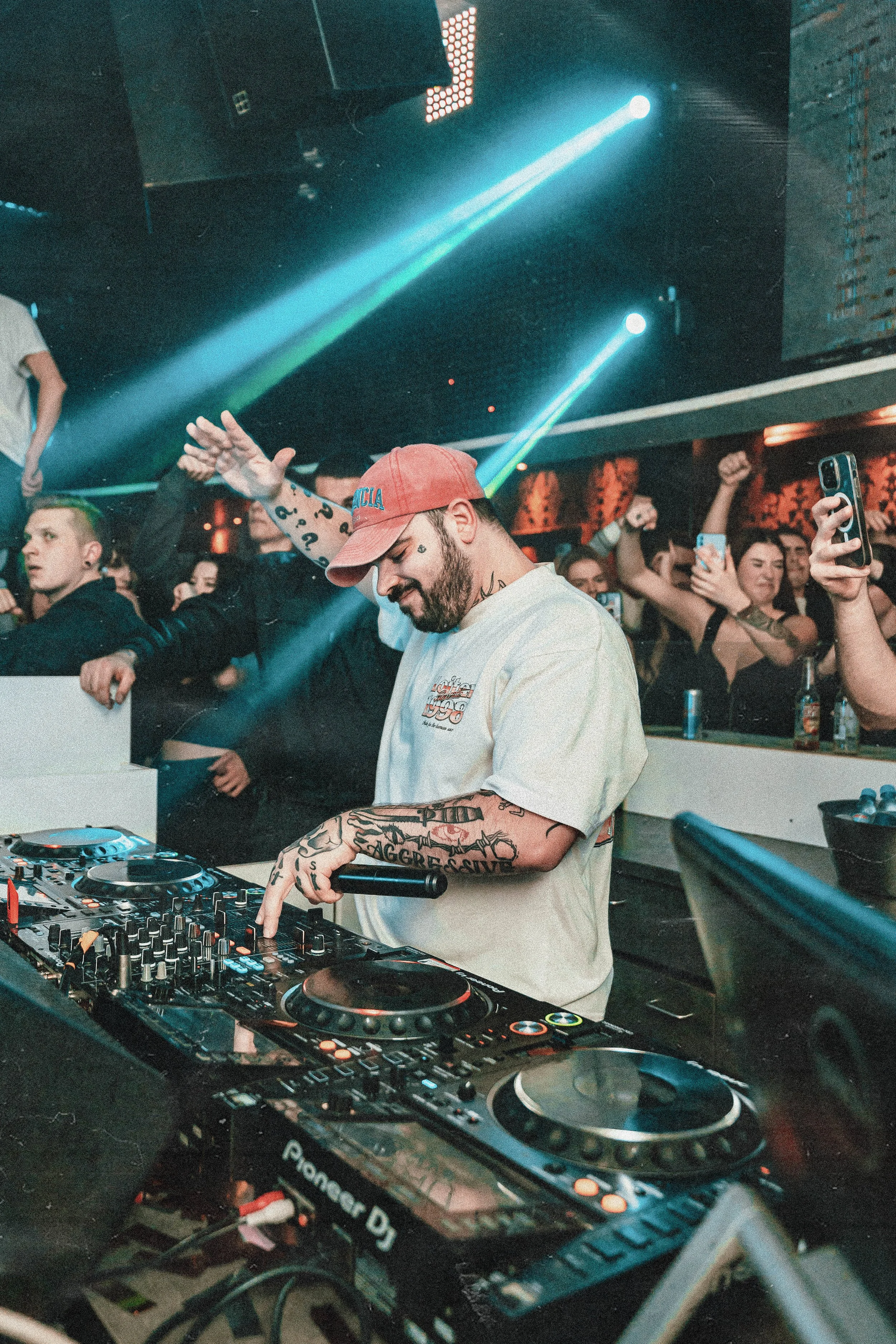 DJ spielt Musik hinter einem Mischpult, umgeben von tanzenden Party-Gästen, mit Laserscheinwerfern im Hintergrund.