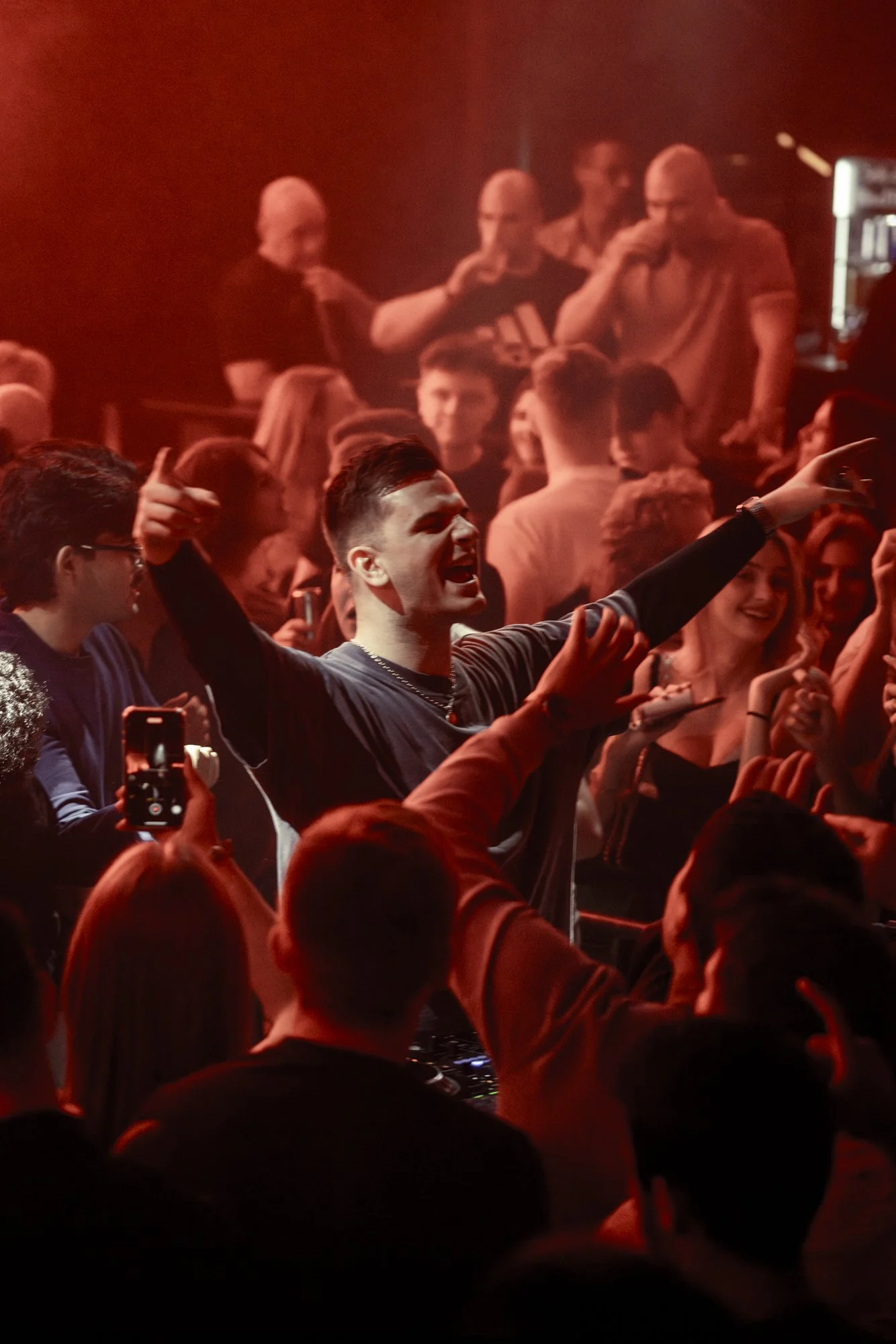 Eine Gruppe von Menschen tanzt und feiert in einem dunklen, rotdurchfluteten Raum, mit einem Jungen in der Mitte, der enthusiastisch singt oder schreit, umgeben von lachenden und jubelnden Personen.