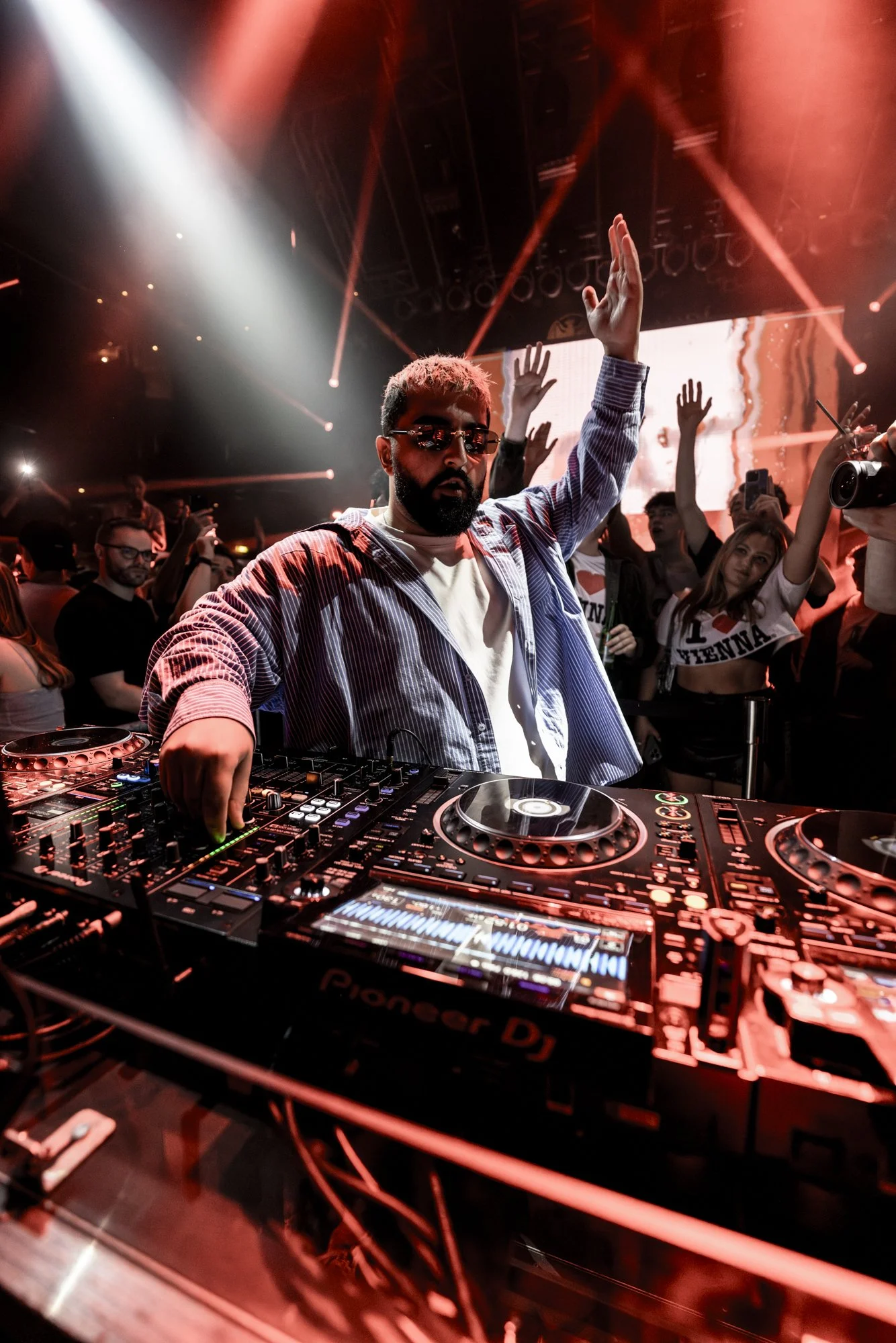 DJ mit Sonnenbrille beim Auflegen in einer Club-Umgebung mit tanzenden Menschen im Hintergrund, rotes Licht und Laserstrahlen.