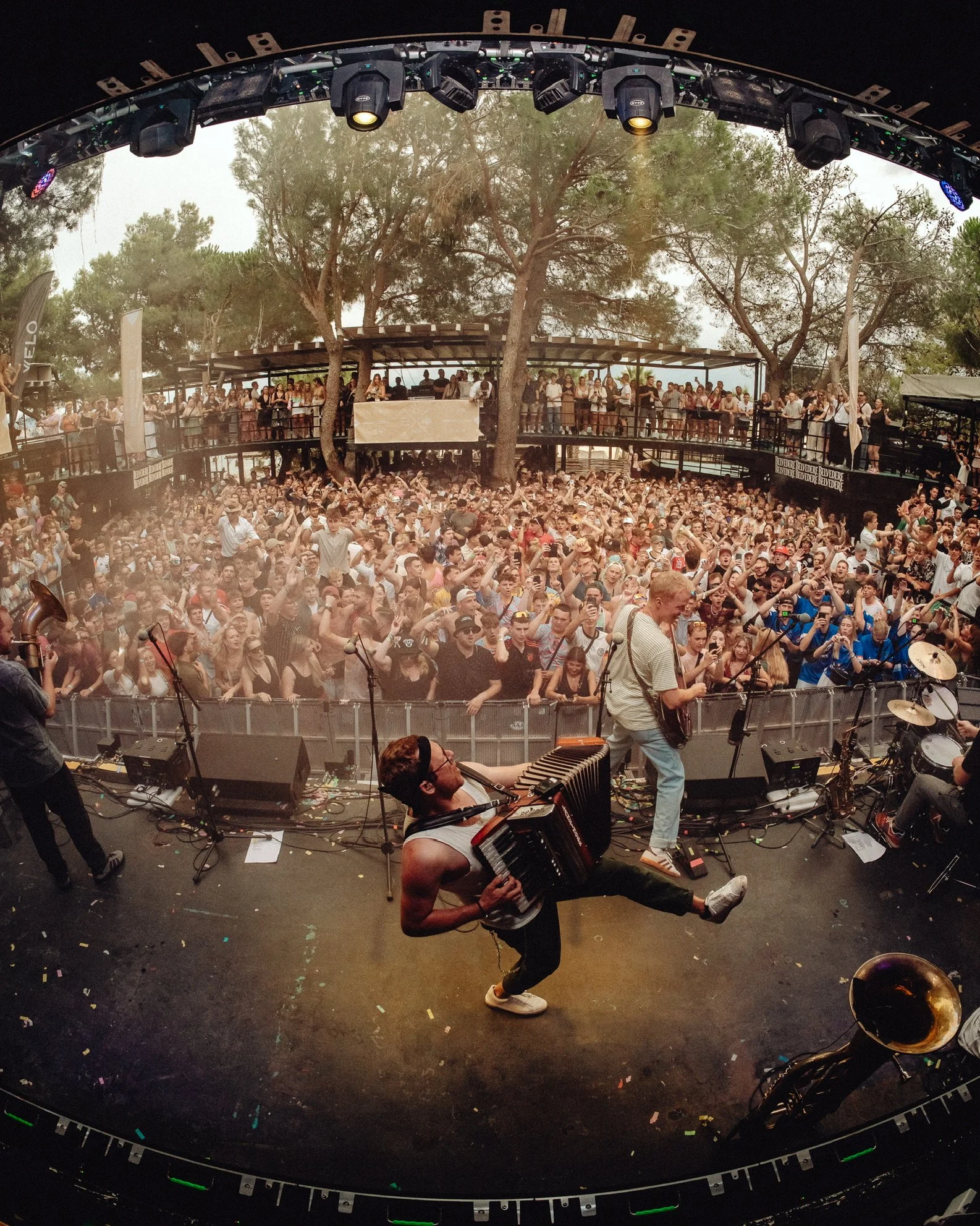 Auftritt auf einer Open-Air-Bühne mit einem Musiker, der ein Keyboard und ein Akkordeon spielt, während eine große Menschenmenge tanzt und zuschaut, umgeben von Bäumen.