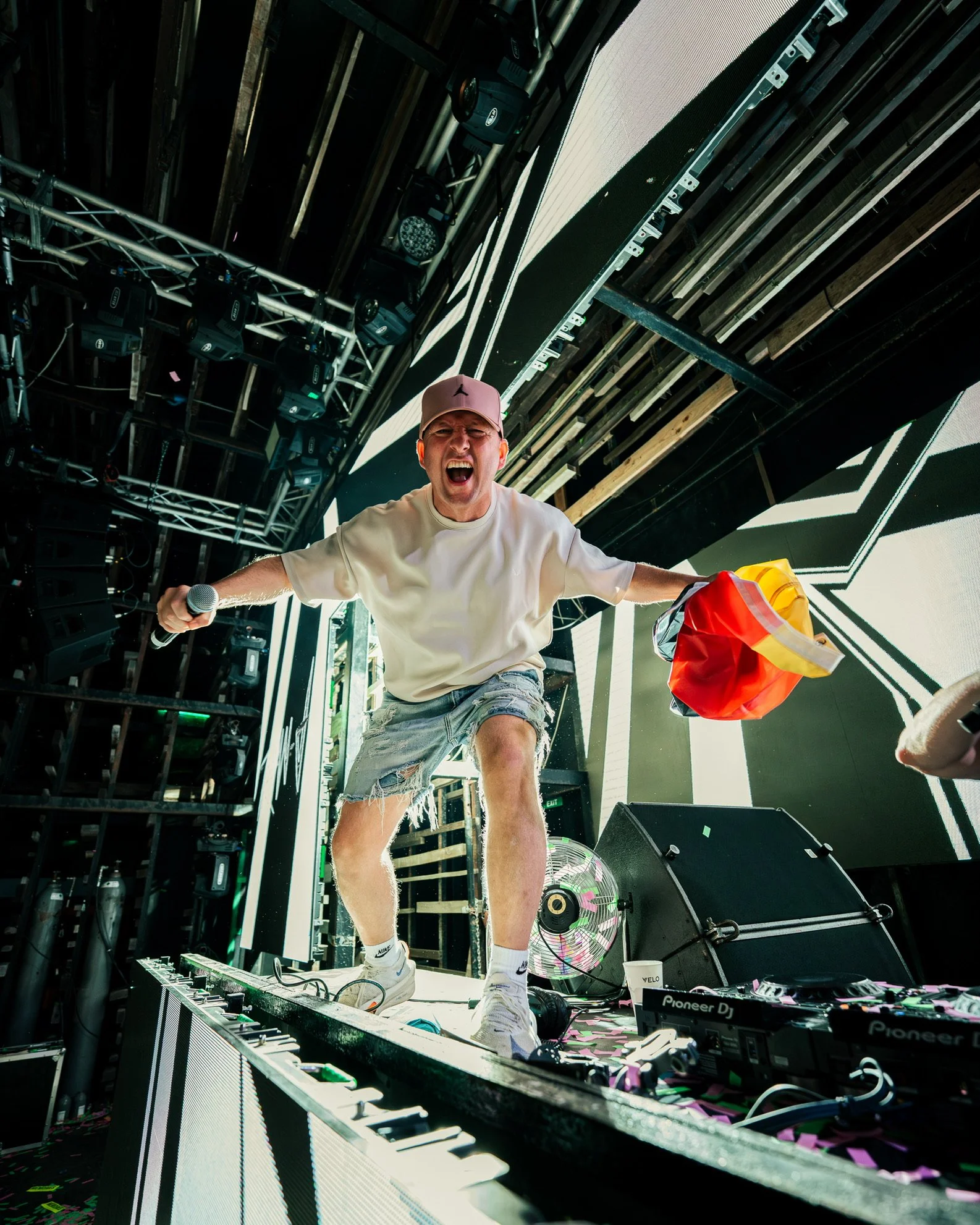 DJ auf der Bühne bei einem Musikfestival, schreit mit einem Mikrofon in der Hand, trägt einen beige Hut, weiße Hemd und zerrissene Jeans, davor DJ-Equipment, bunte Verpackungen und ein Fan, im Hintergrund Lichter und Bühnenaufbau.