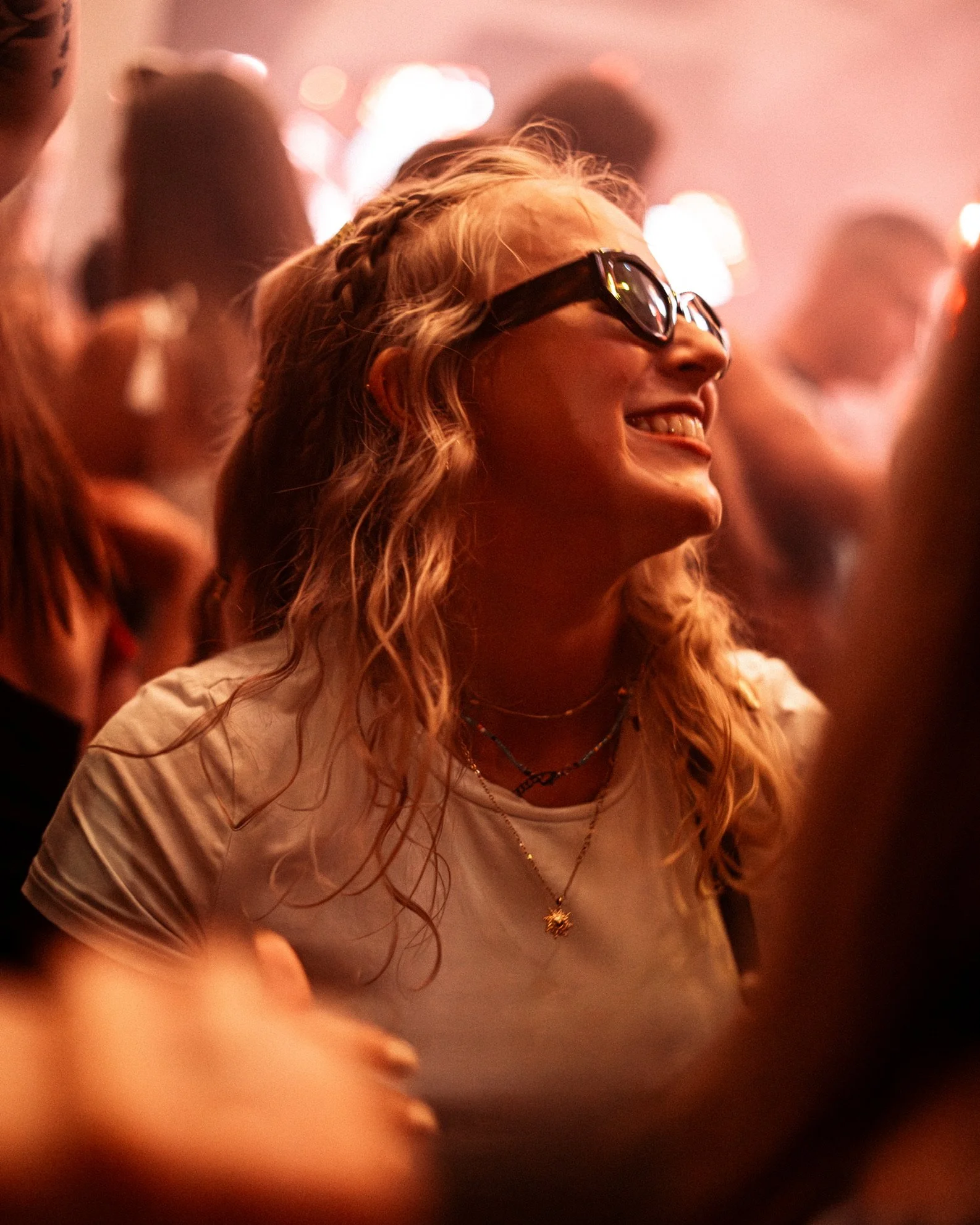 Eine lachende junge Frau mit lockigem Haar, Sonnenbrille und mehreren Halsketten auf einem Konzert oder Festival, umgeben von anderen Menschen im gleichen Umfeld.