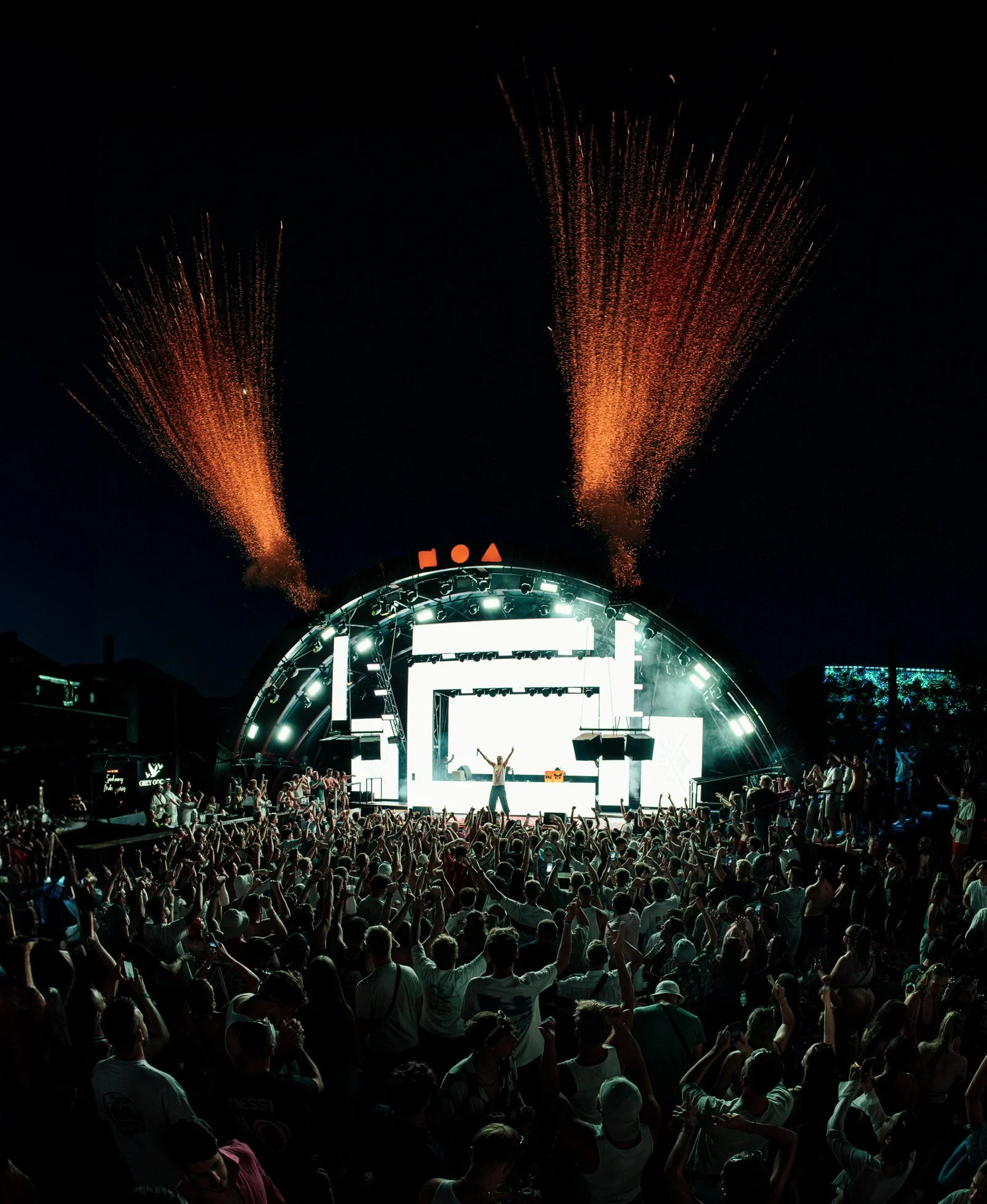 Eine große Menschenmenge bei einem nächtlichen Open-Air-Musikfestival vor einer Bühne, auf der ein DJ steht, mit Feuerwerk am Himmel.
