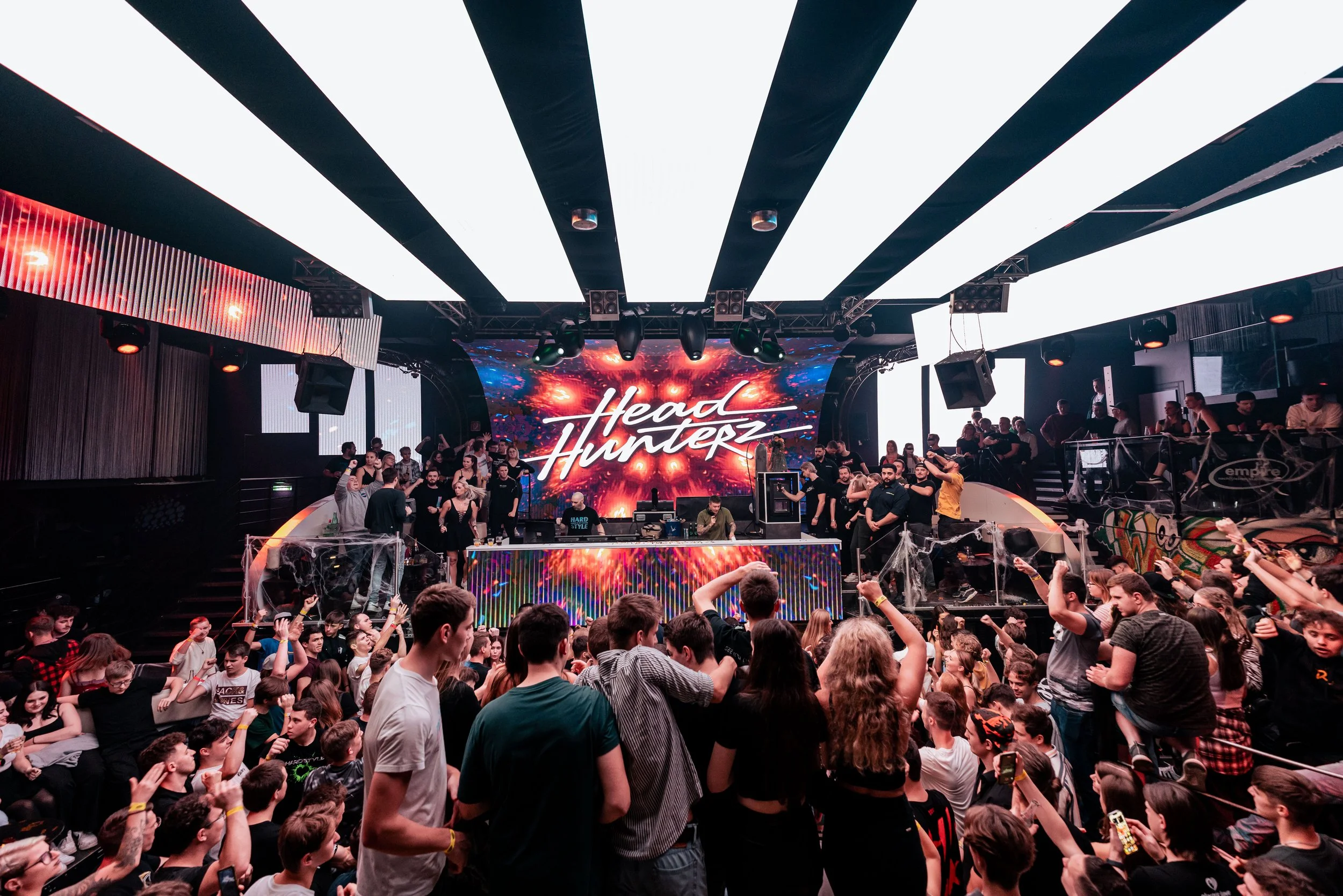 Menschen tanzen und feiern auf einer Party im Club mit einer großen Bühne, einem DJ und einer zentralen LED-Wand, auf der ‚Head Hunterz‘ steht.