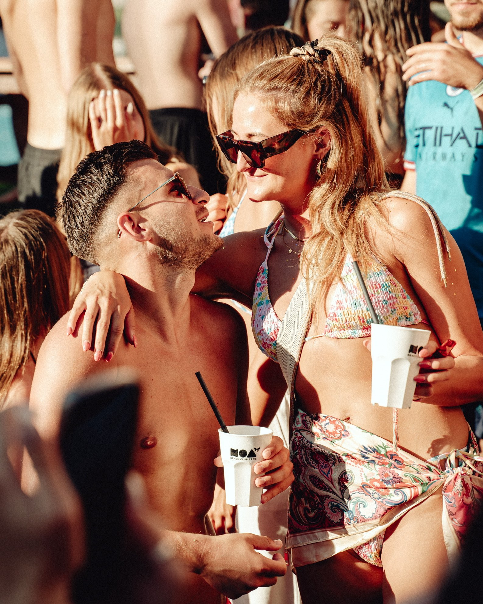 Zwei junge Erwachsene, ein Mann und eine Frau, stehen sich nahe bei einer Party im Freien, lachen und schauen sich an, während sie Getränke in Bechern halten.