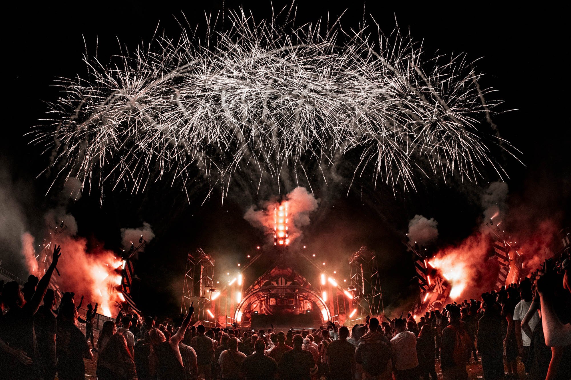 Feuerwerk über einem Konzert mit einer Menschenmenge bei Nacht, mit roten Lichtern und Rauch.