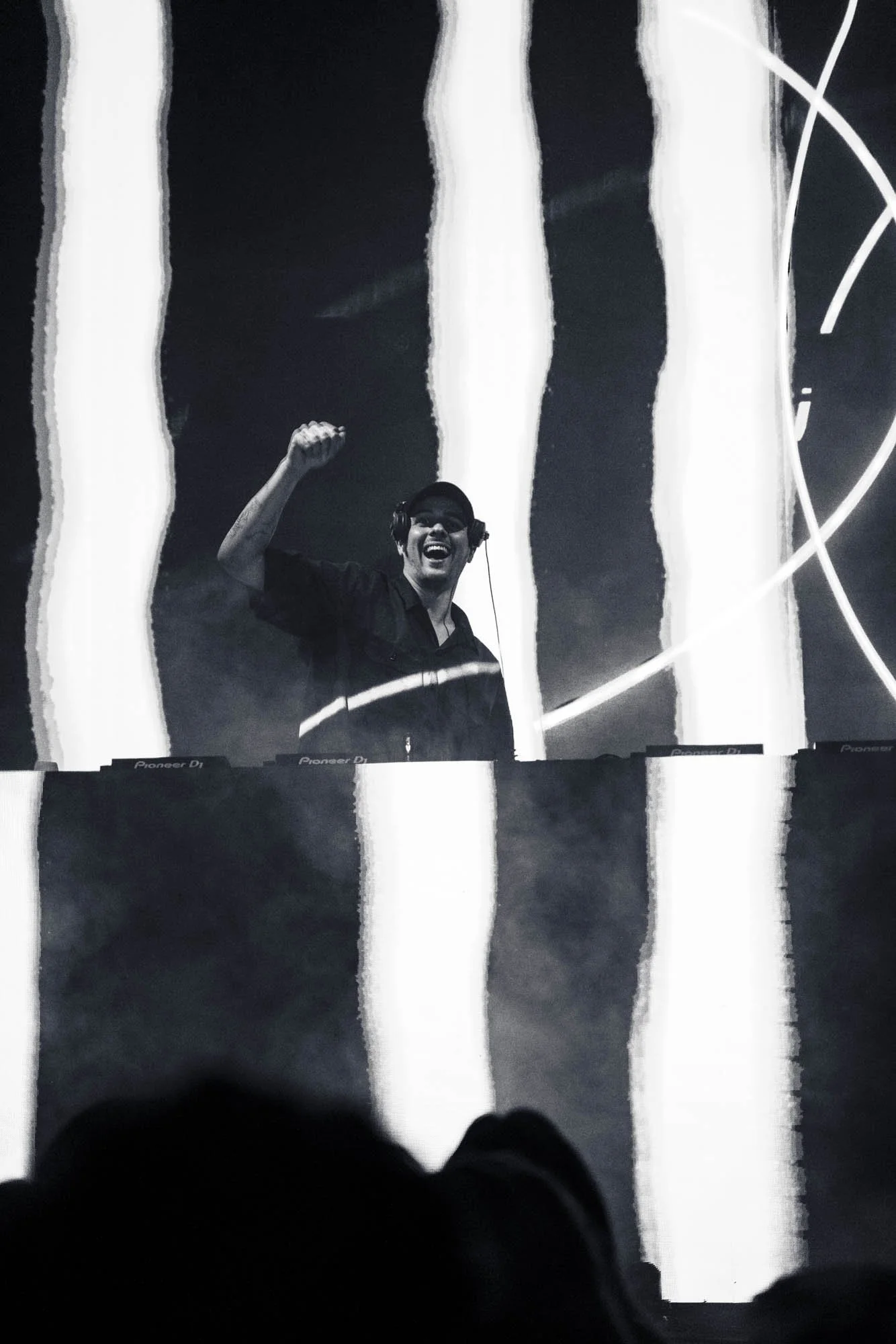 Ein DJ mit Headset, der auf der Bühne hinter einem DJ-Pult steht, jubelt mit erhobener Faust. Der Hintergrund zeigt abstrakte, vertikale weiße Lichtstreifen auf einer dunklen Fläche.