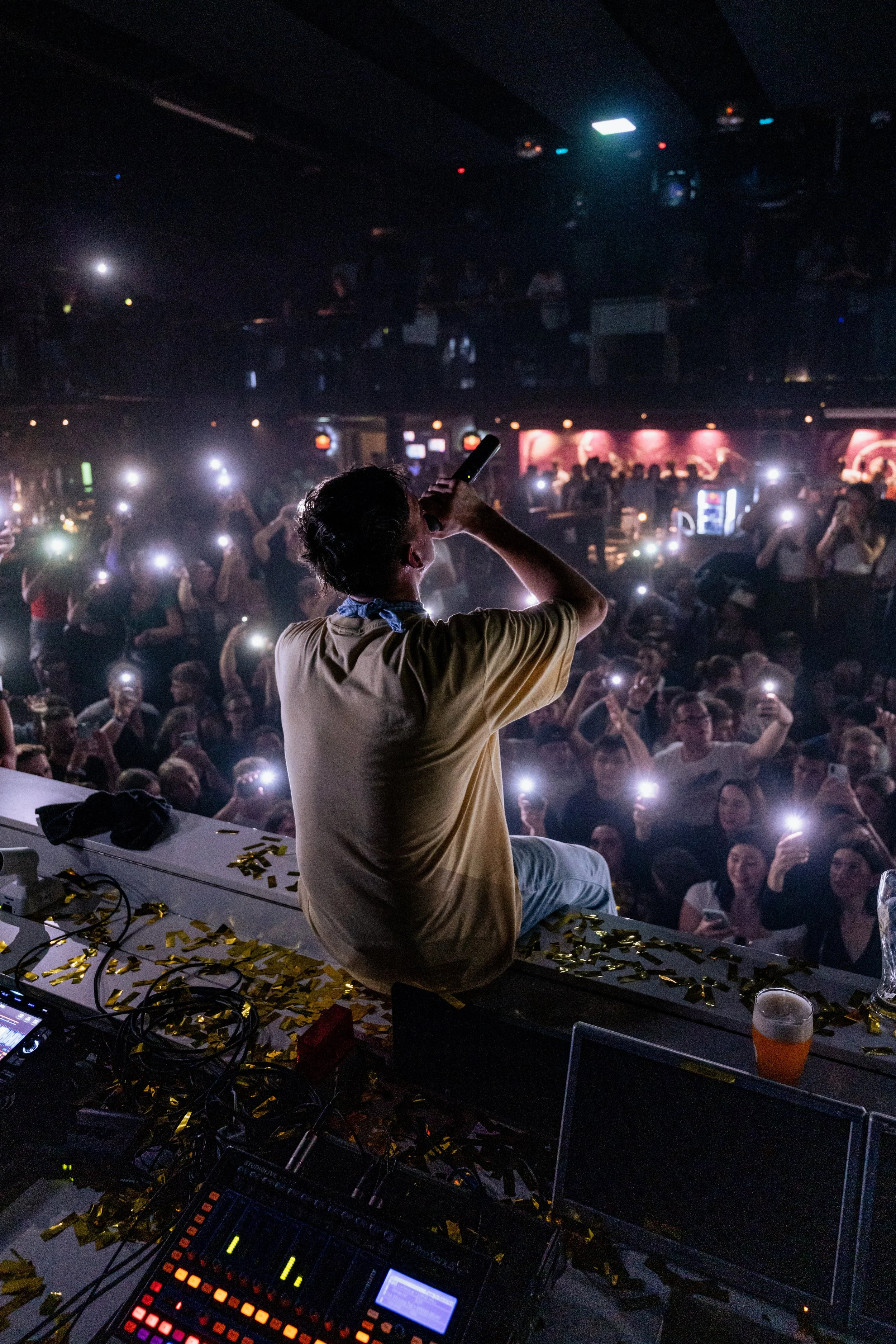 DJ auf einer Bühne während eines Konzerts oder einer Party, blickt auf eine große Menge von Menschen, die mit ihren Mobiltelefonen Lichter blitzen und Fotos machen. Der Raum ist dunkel, mit einigen bunten Lichtern im Hintergrund und Konfetti auf der 