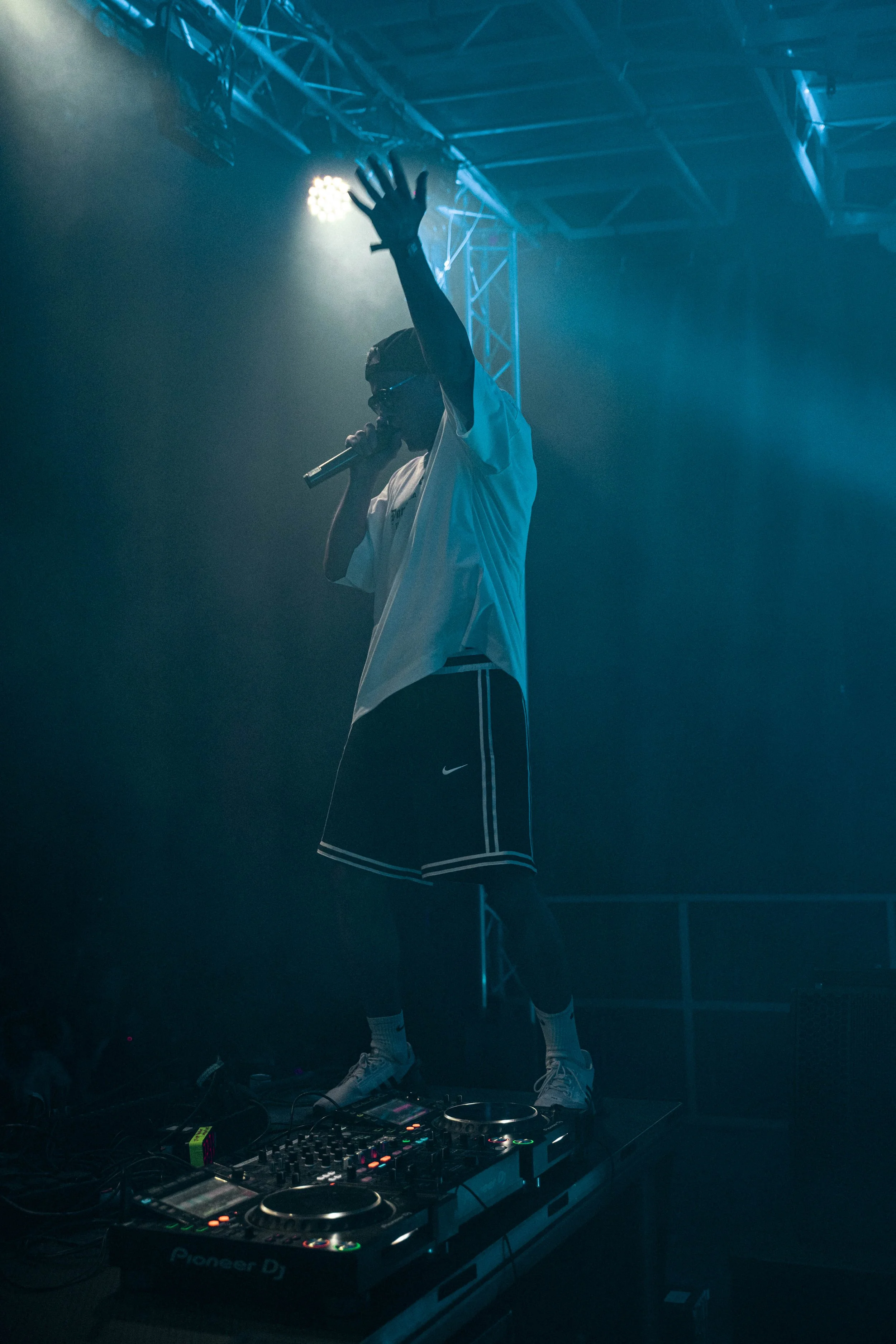 Ein DJ mit Kopfhörern, weißen T-Shirt, schwarzen Shorts und Turnschuhen steht auf einem DJ-Pult, während er singt und in der Hand ein Mikrofon hält, bei einer Nachtveranstaltung mit blauer und dunkler Beleuchtung.