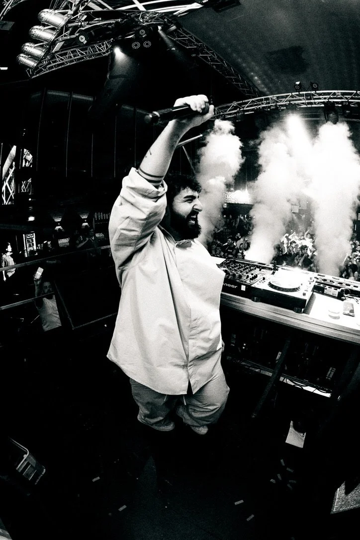 Mann mit Mikrofon bei einer DJ-Performance in einem Club, umgeben von Rauch und einem Publikum.