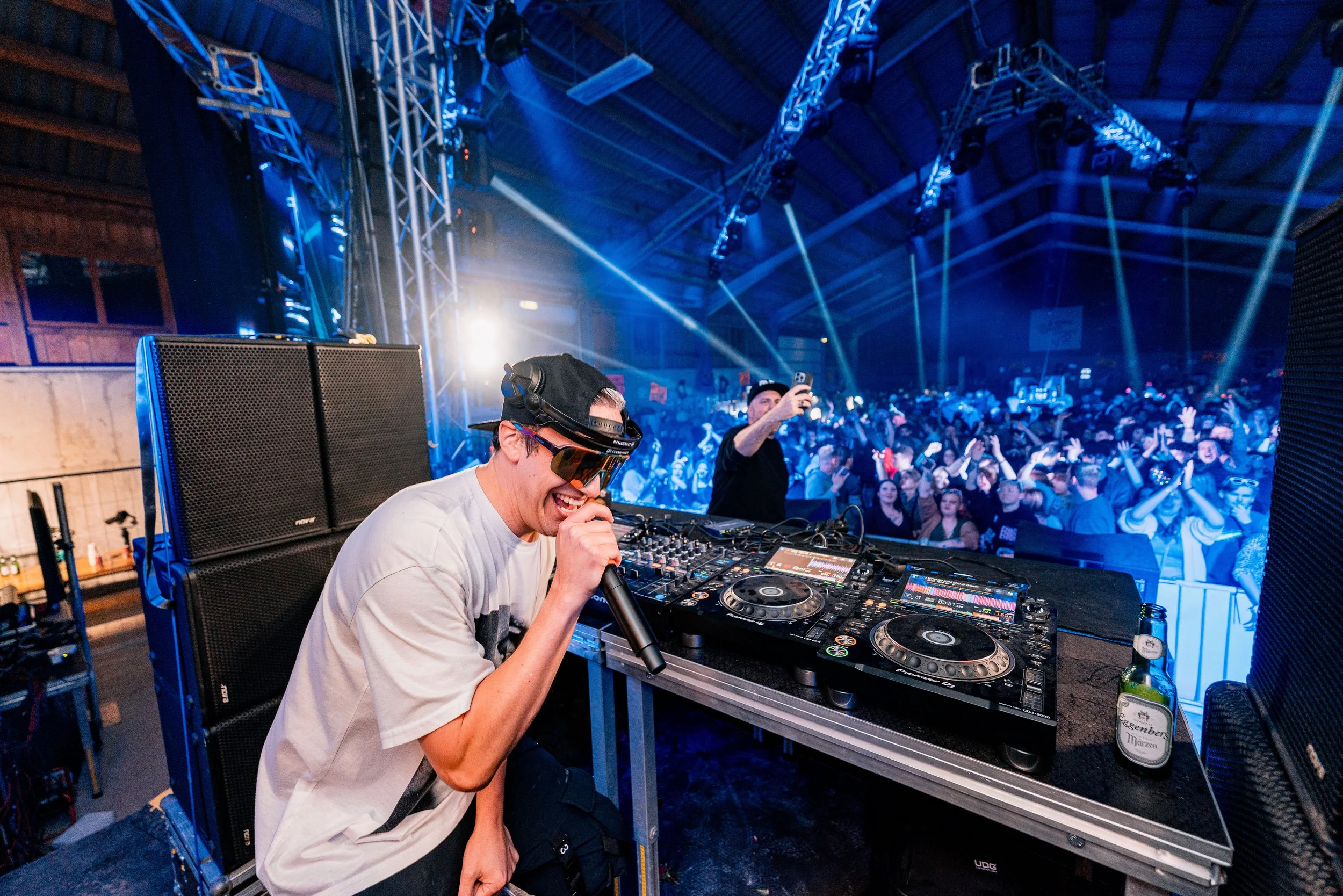 DJ auf der Bühne in einer rockigen oder elektronischen Musikveranstaltung, mit Publikum und Lichtshows im Hintergrund.