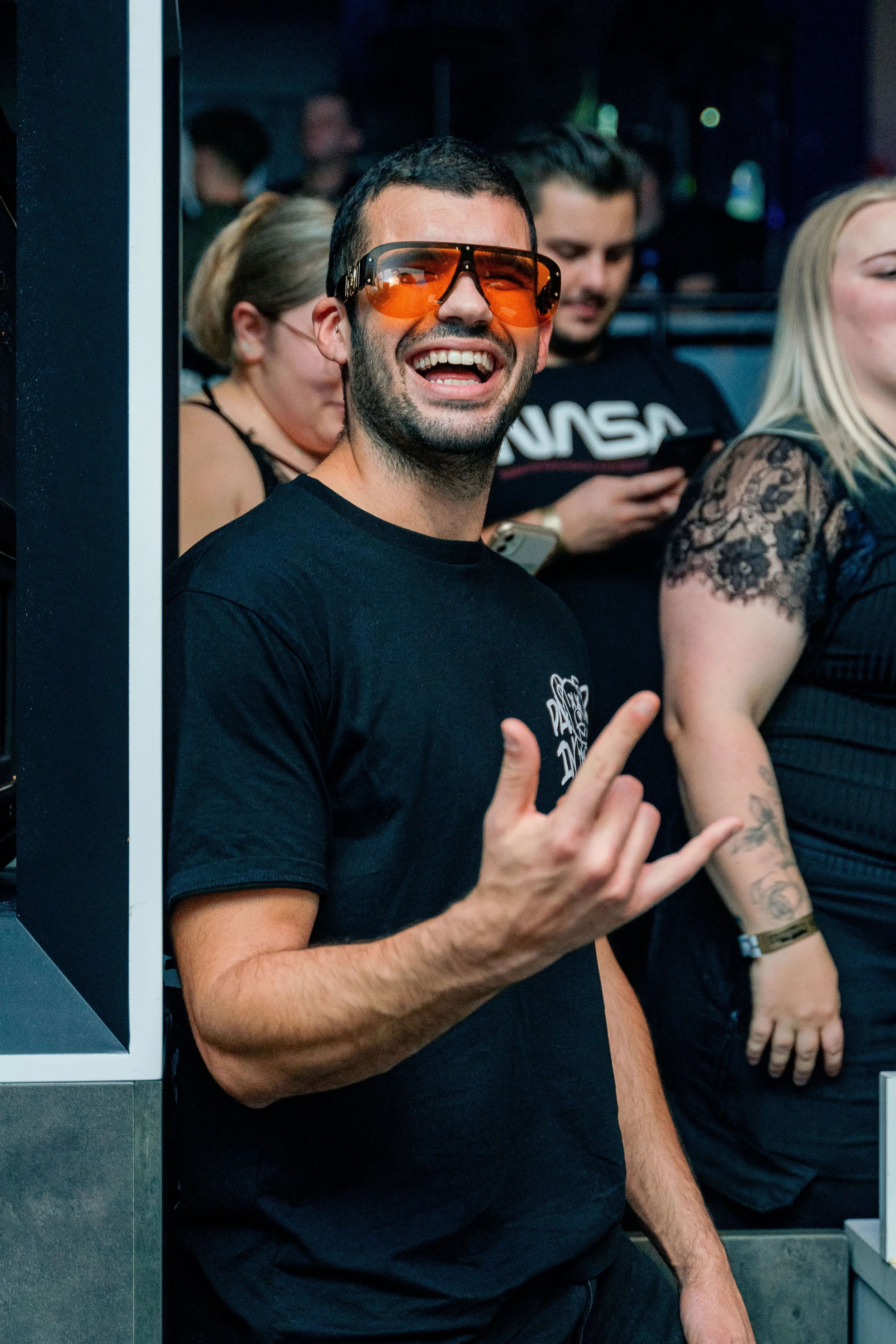 Ein lachender junger Mann mit dunklem Haar, Brille mit orangefarbenen Gläsern, trägt ein schwarzes T-Shirt und zeigt das Handzeichen für Heavy Metal in einer Menge bei einer Veranstaltung.