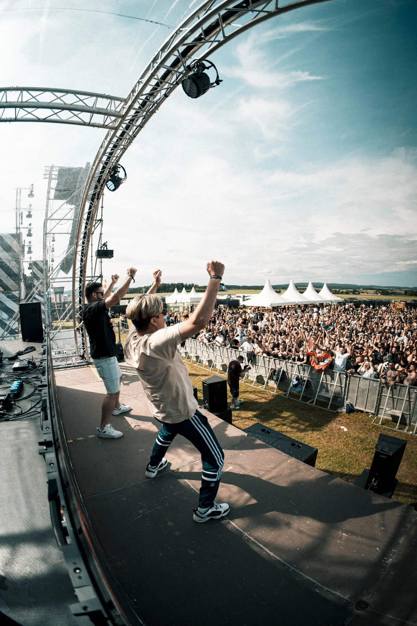 Auf einer Open-Air-Bühne tanzen zwei Menschen vor einem großen Publikum bei einem Musikfestival, Sonnenwetter, Himmel mit Wolken, weiße Zelte im Hintergrund.