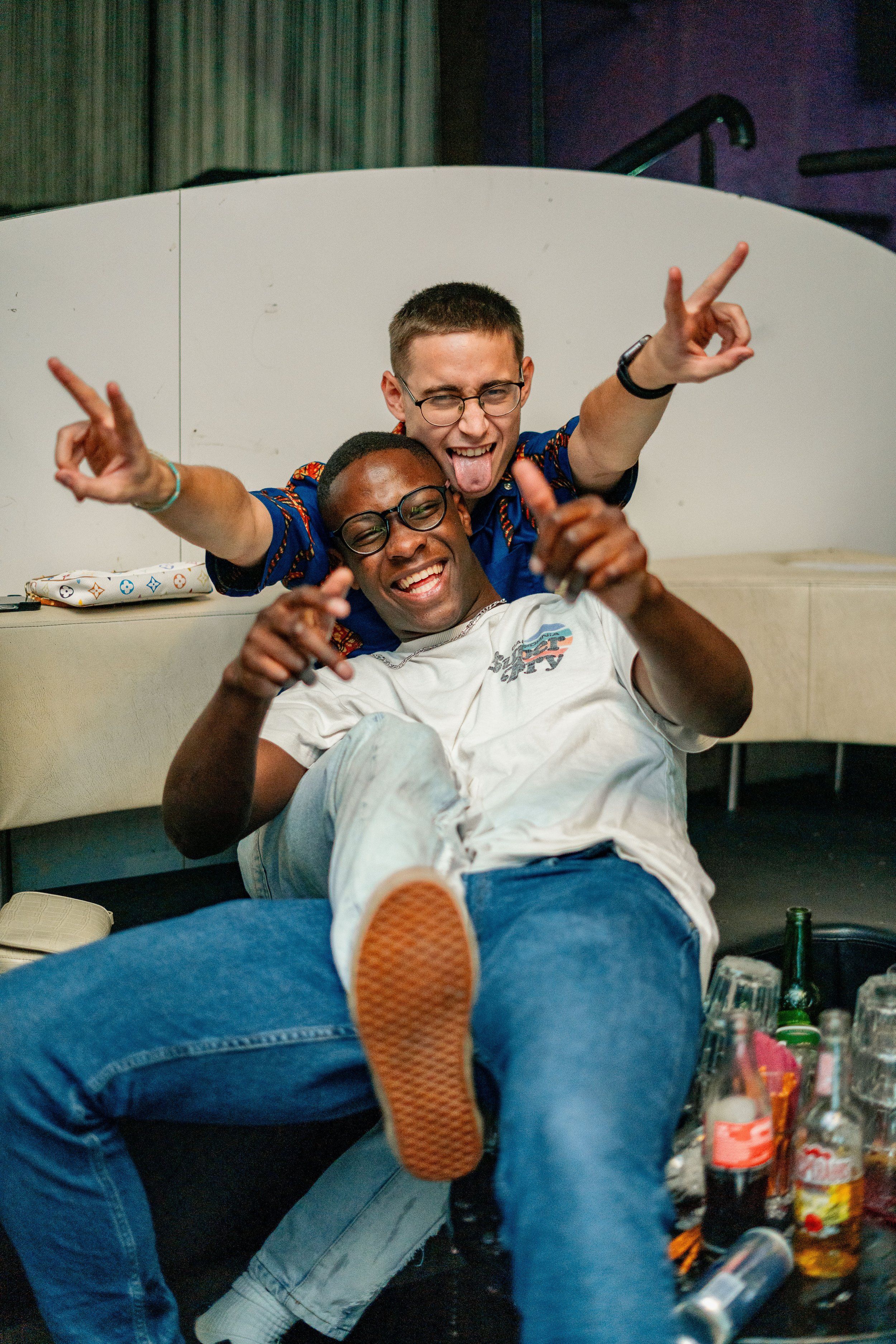 Zwei lachende Männer, einer sitzt auf dem Boden und der andere sitzt auf seinem Schoß, in einer Bar, umgeben von leeren Flaschen und Gläsern, feiern und haben Spaß.