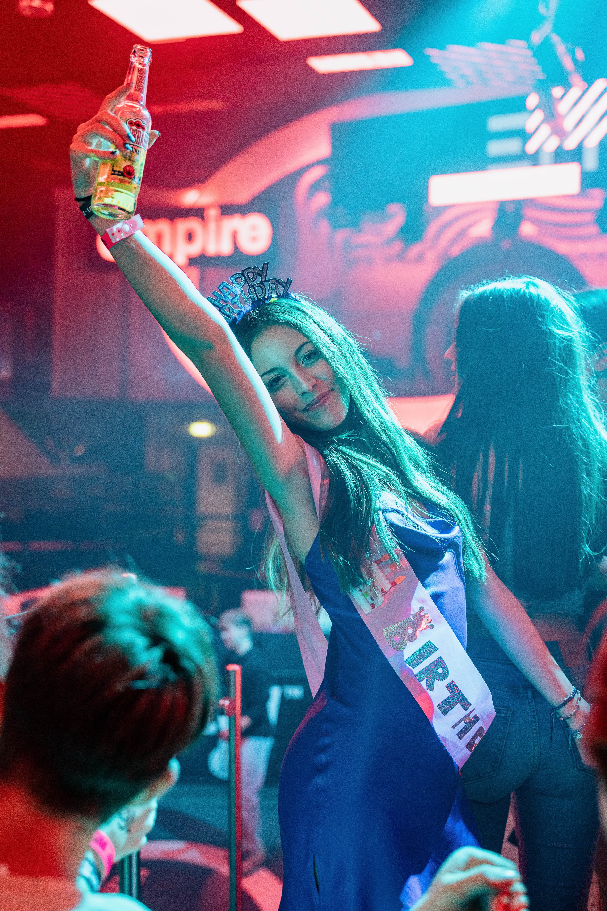 Junge Frau trägt eine „Happy Birthday“-Krawatte und eine Krone, feiert in einem Club, hält eine Bierflasche hoch, umgeben von Leuten, mit bunt beleuchteter Atmosphäre.