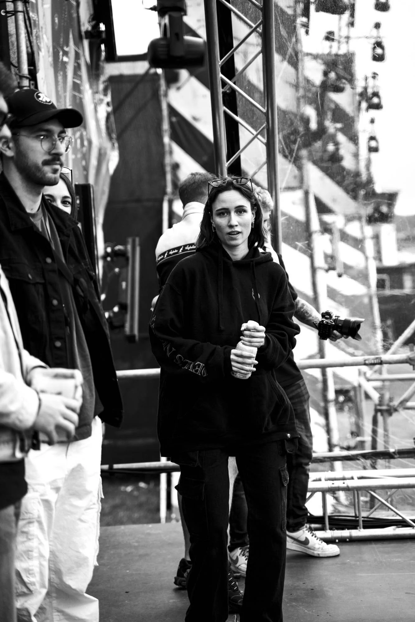 Menschen auf einer Bühne oder bei einem Event, schwarz-weiß. Die zentrale Frau trägt eine schwarze Kapuzenjacke, hält ihre Hände zusammen und schaut in die Kamera. Weitere Personen stehen links und im Hintergrund, einige mit Getränken in der Hand. Bü