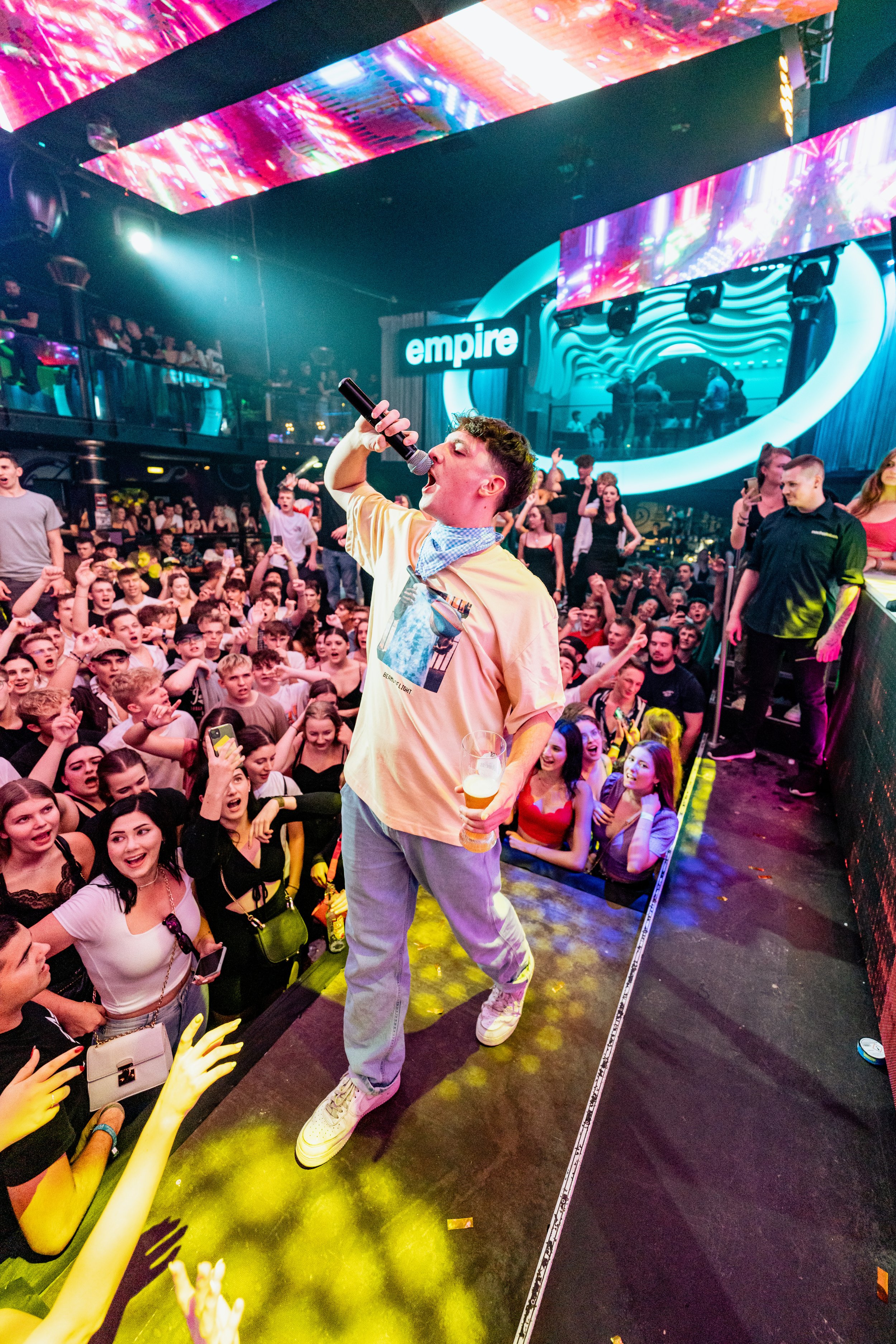 Junger Sänger in einem Club mit vielen tanzenden Menschen im Hintergrund, Bühne und die Silhouette des DJs mit dem Empire-Logo im Hintergrund.