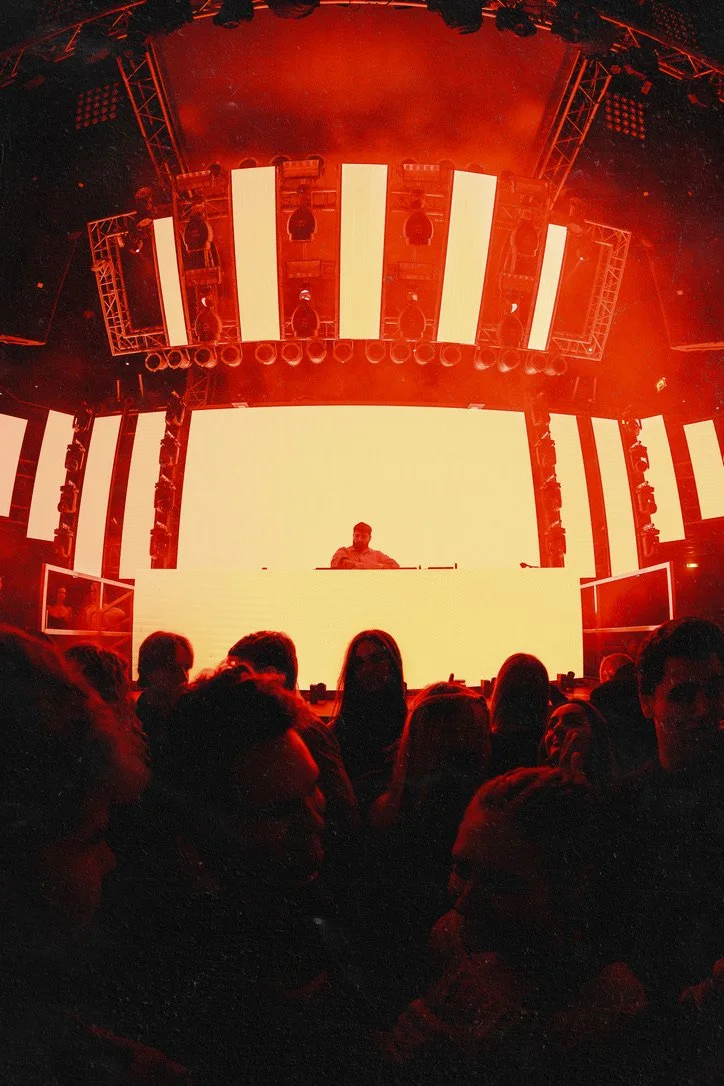 Menschenmenge vor einer großen, beleuchteten Bühne mit einem DJ oder Musiker im Mittelpunkt bei einem Konzert oder Festival.