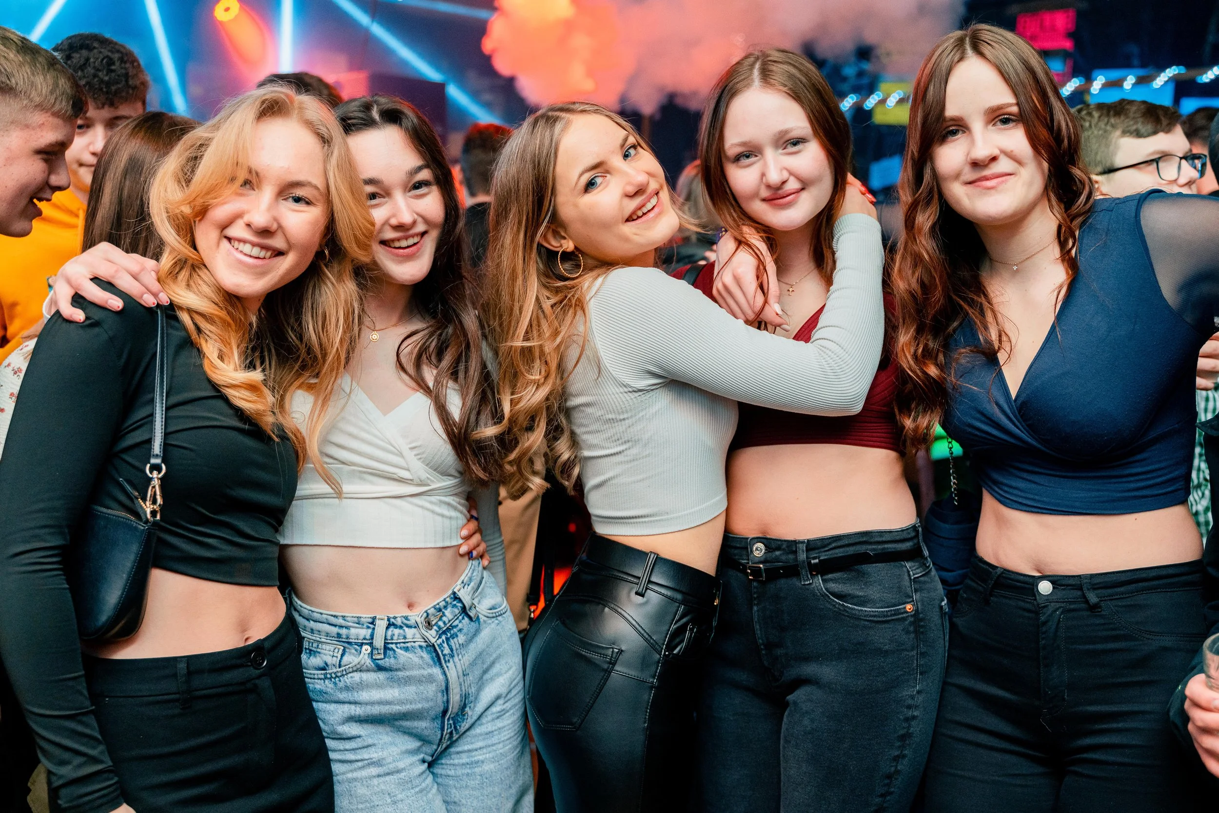 Fünf Jugendliche Frauen, die auf einer Party lächeln und Fotos machen, umgeben von weiteren feiernden Menschen und bunter Lichtshow.