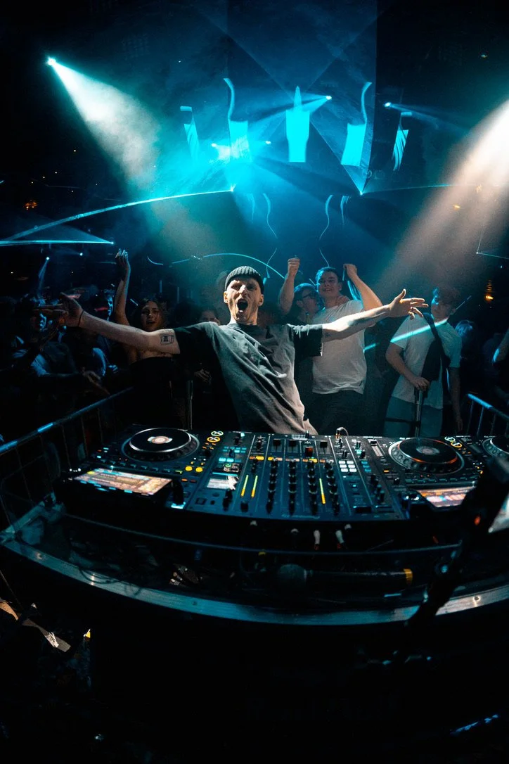 DJ steht vor seinem Mischpult auf einer Party, umgeben von tanzenden Menschen, mit buntem Licht und Nebel im Hintergrund.