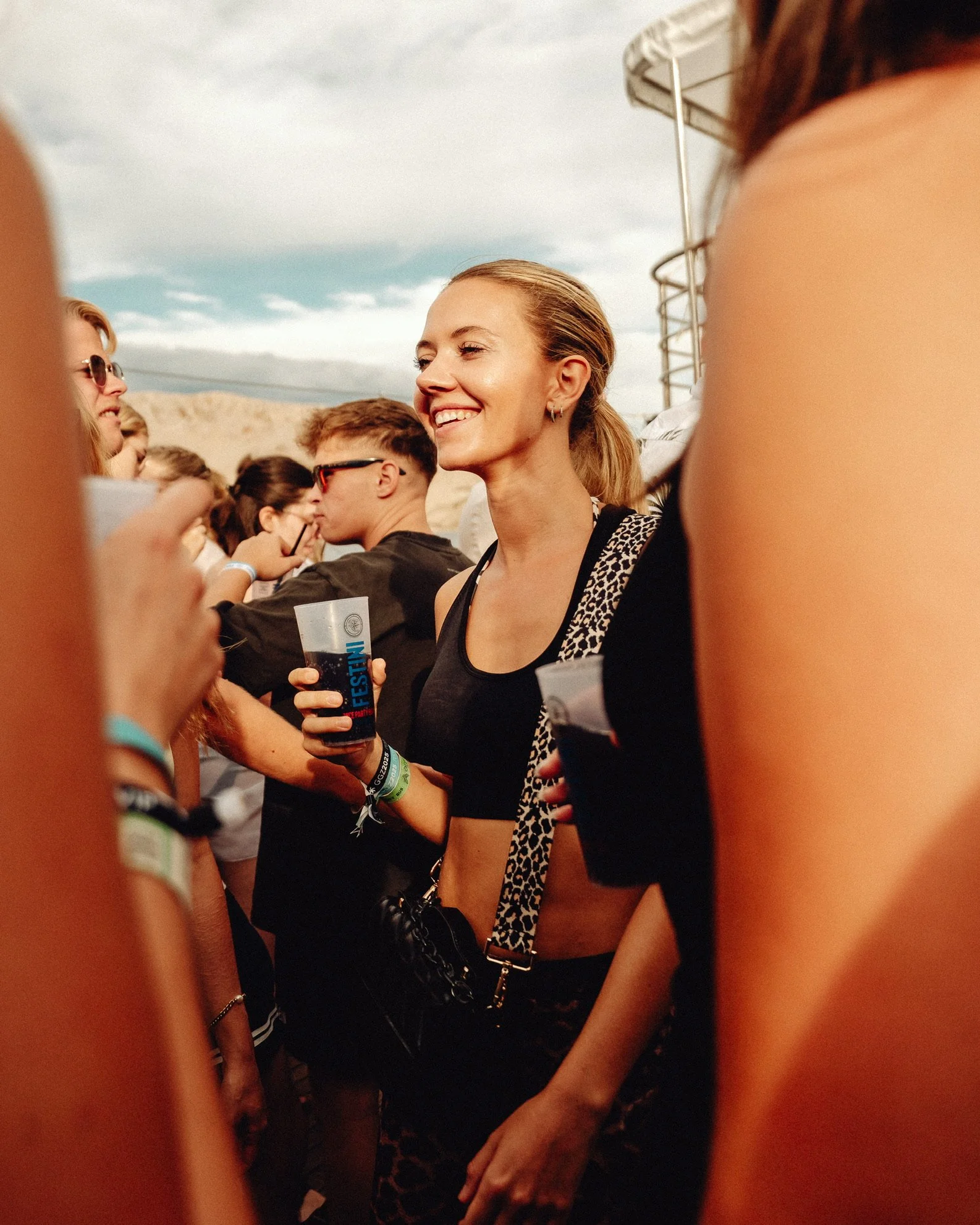 Eine junge Frau bei einem Musikfestival, die ein Getränk hält, umgeben von anderen Festivalbesuchern im Freien.