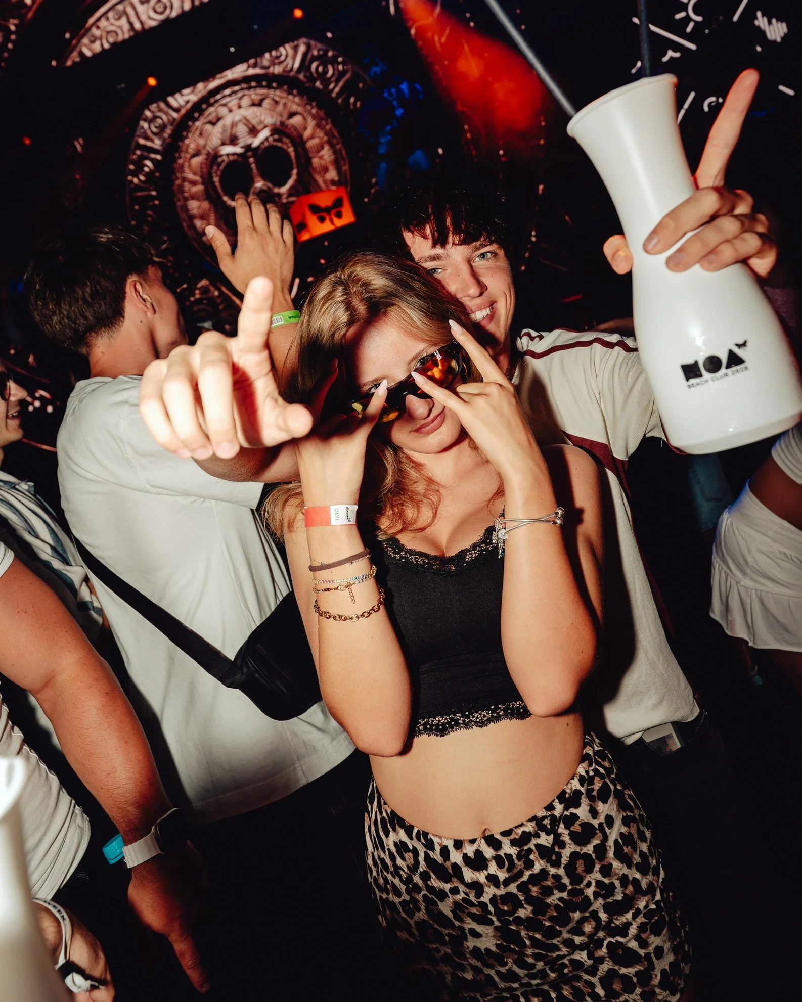 Menschen beim Feiern auf einer Party, einige tragen Sonnenbrillen, eine Person hält eine Vase, und im Hintergrund eine Lasershow.